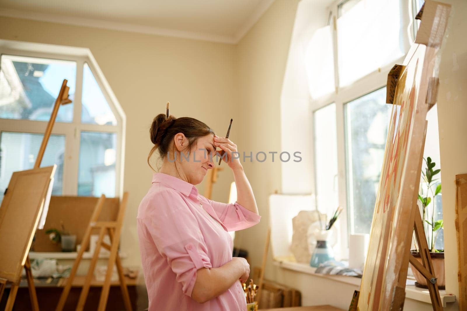 Caucasian woman artist working on a painting in bright daylight studio. Happy artist draws an art project with paints and a brush in the workshop. Hobby. Artist at work. Creative profession by Tomashevska