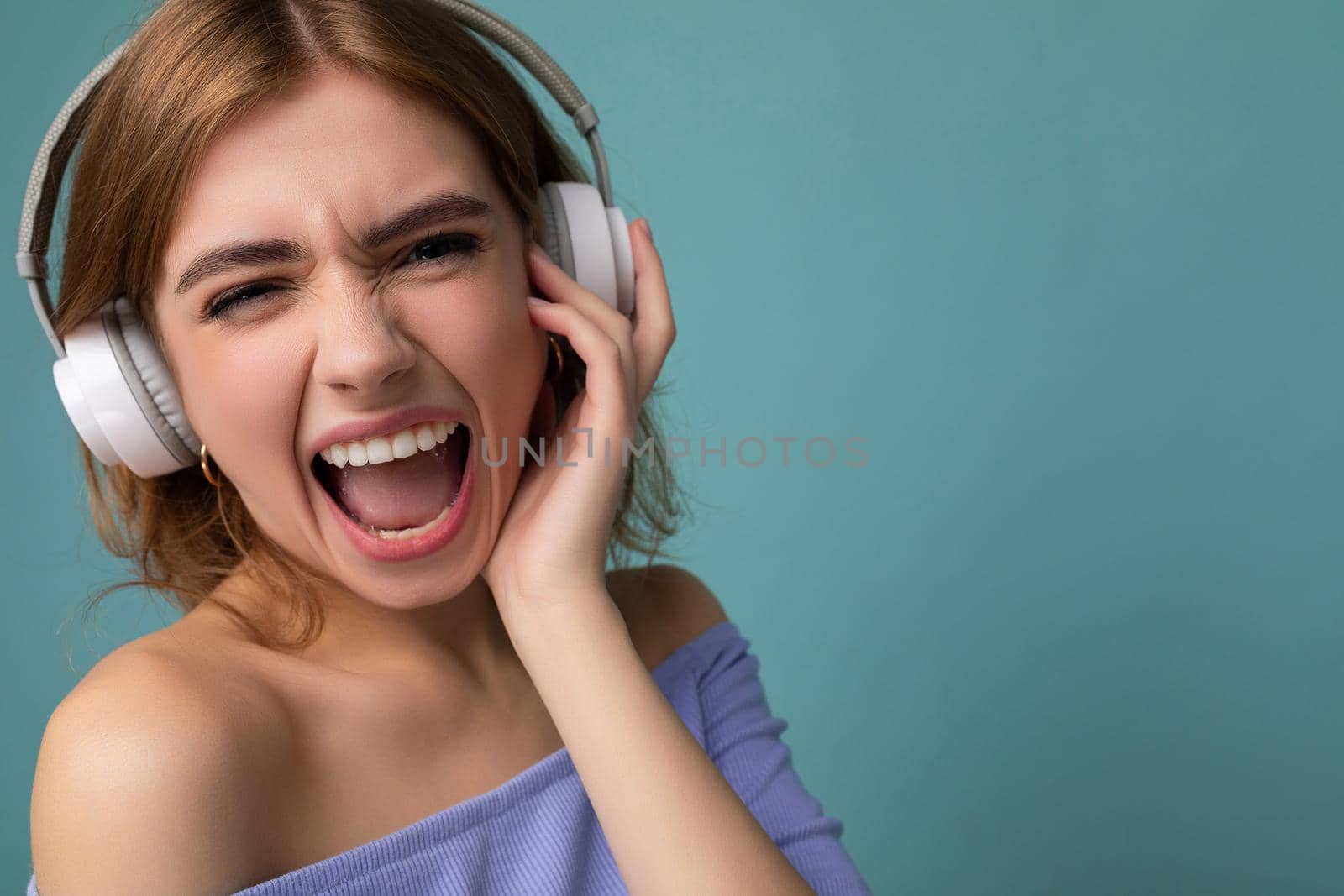 Closeup portrait of attractive emotional young blonde woman wearing blue crop top isolated on blue background wearing white wireless bluetooth headphones listening to cool music and having fun looking at camera.