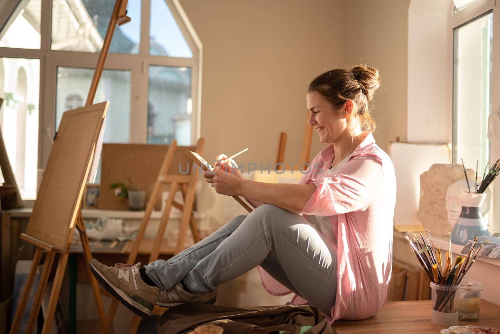 Cute woman paints on canvas in an art workshop. Artist creating picture. Art school or studio. Work with paints, brushes and easel. Hobby and leisure concept. Woman painter at workspace.