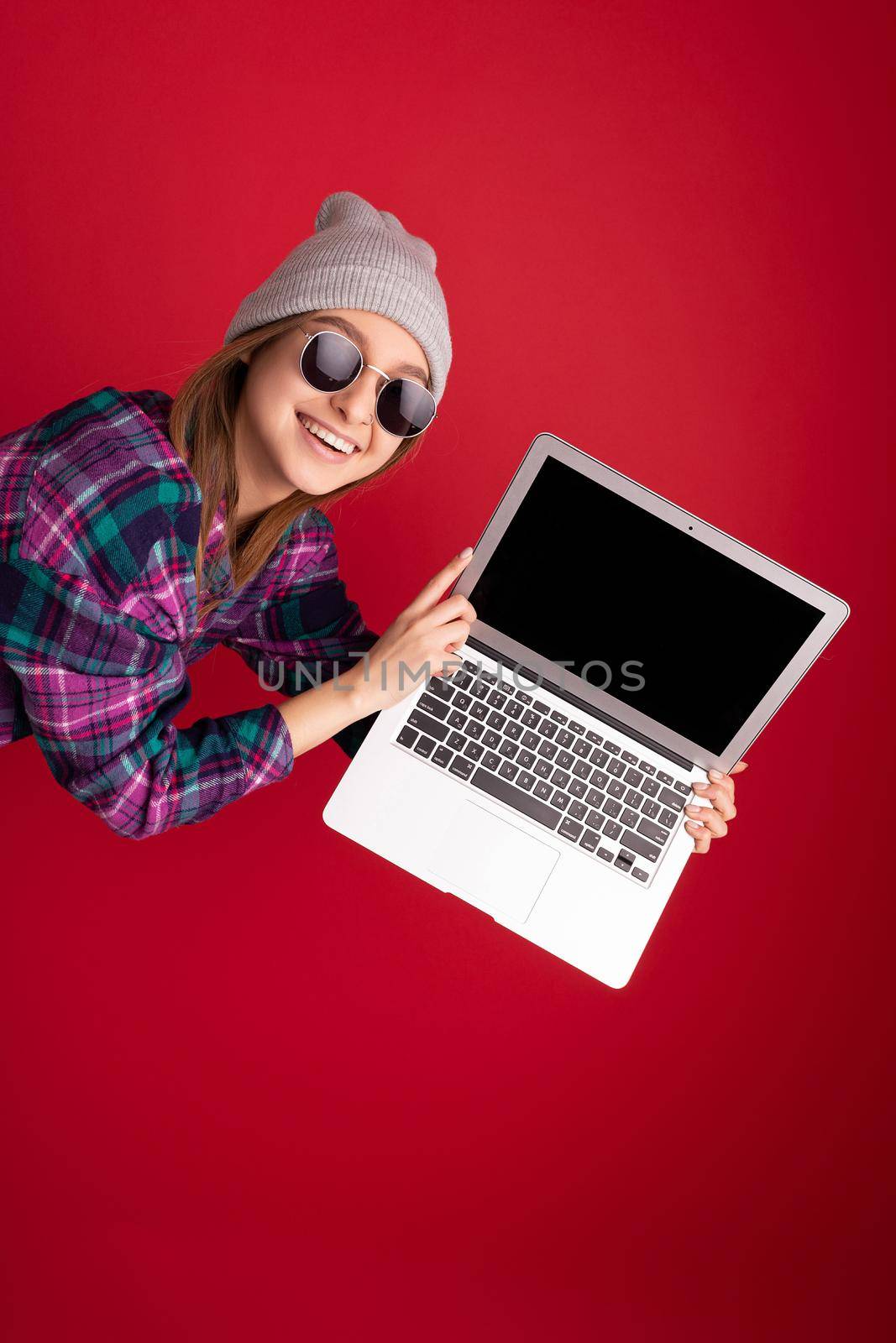 Close-up portrait of crazy mad overjoyed Beautiful smiling happy young woman holding netbook computer looking at camera having fun wearing sunglasses colourful shirt grey hat isolated over red wall background by TRMK