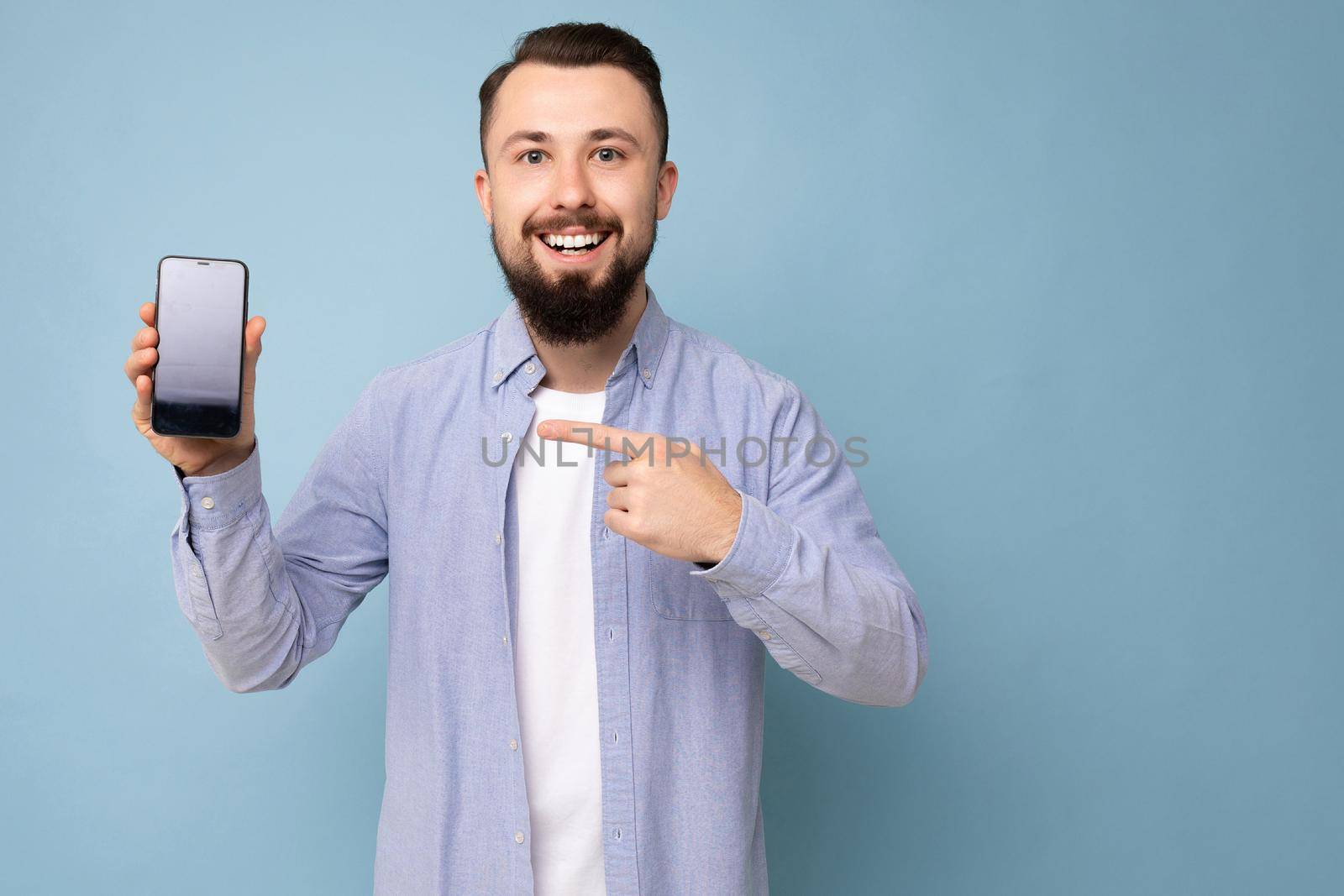 Photo of handsome smiling adult male person good looking wearing casual outfit standing isolated on background with copy space holding smartphone showing phone in hand with empty screen display for mockup pointing at gadjet looking at camera by TRMK