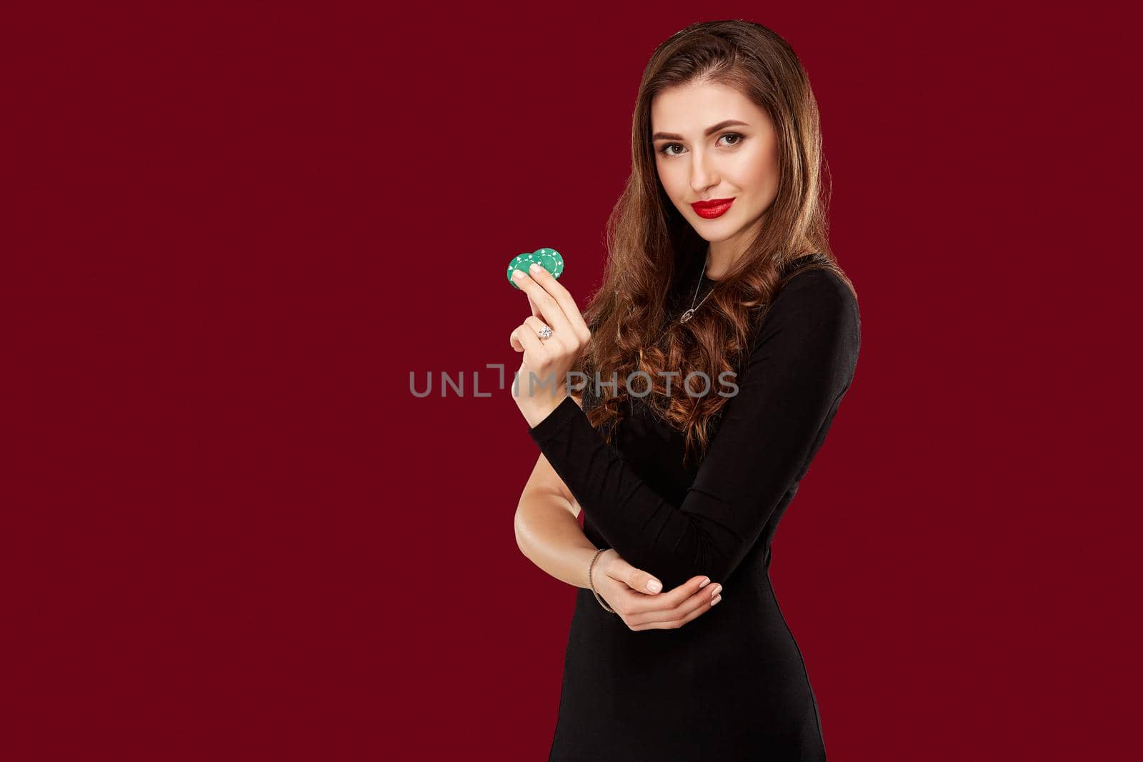Pretty long hair woman in black dress holding chips for gambling in casino. Studio shot on red background. Poker. Two green Chips in Hand