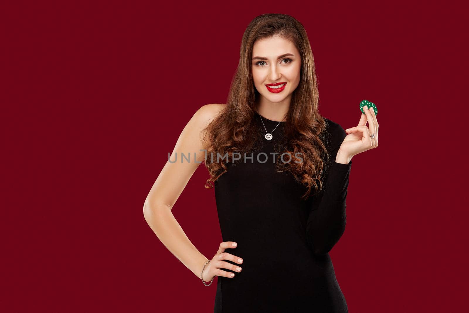 Pretty long hair woman in black dress holding chips for gambling in casino. Studio shot on red background. Poker. Two green Chips in Hand