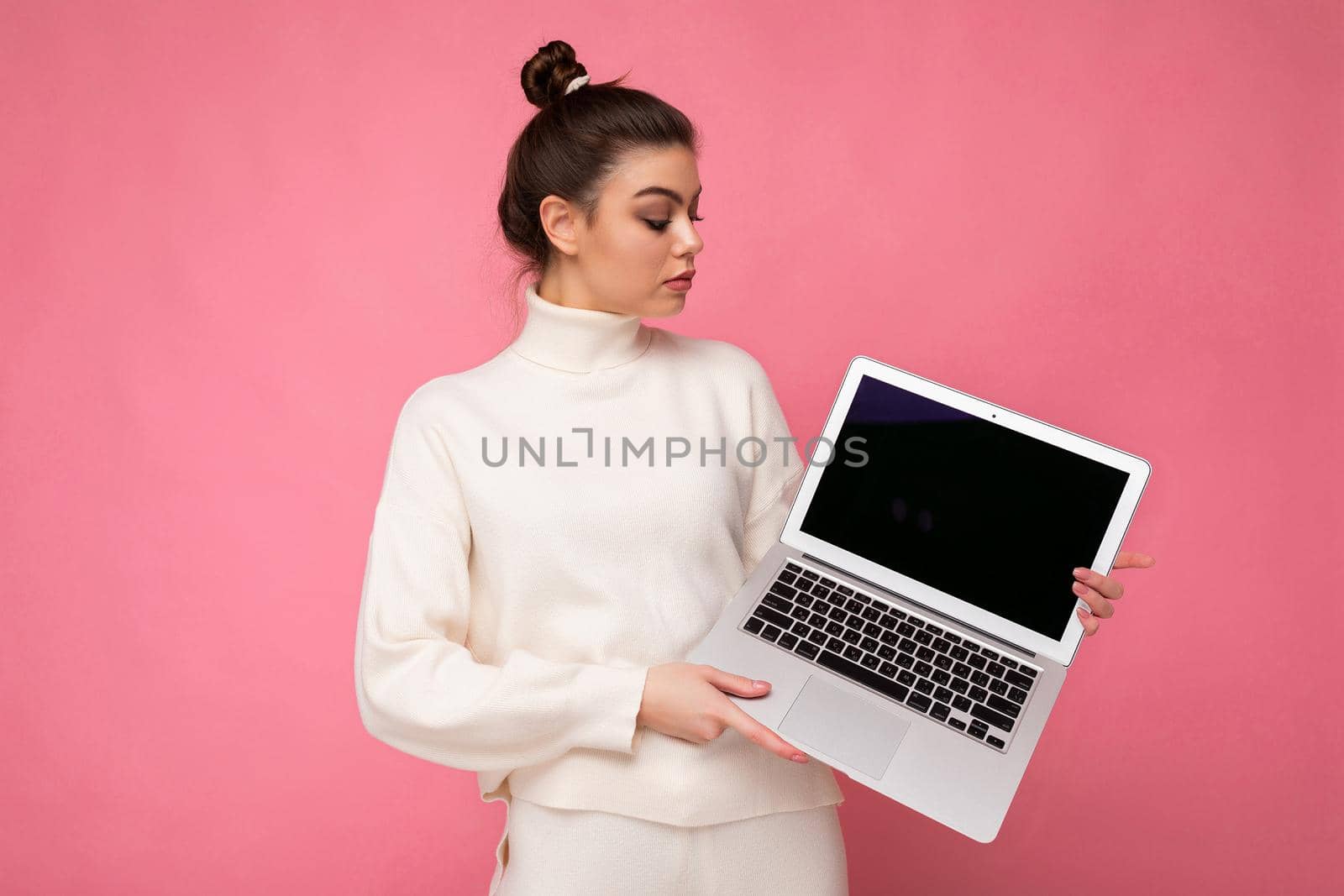 Photo of beautiful woman with gathered brunette hair wearing white sweater holding computer laptop and looking at the netbook isolated over pink wall background.
