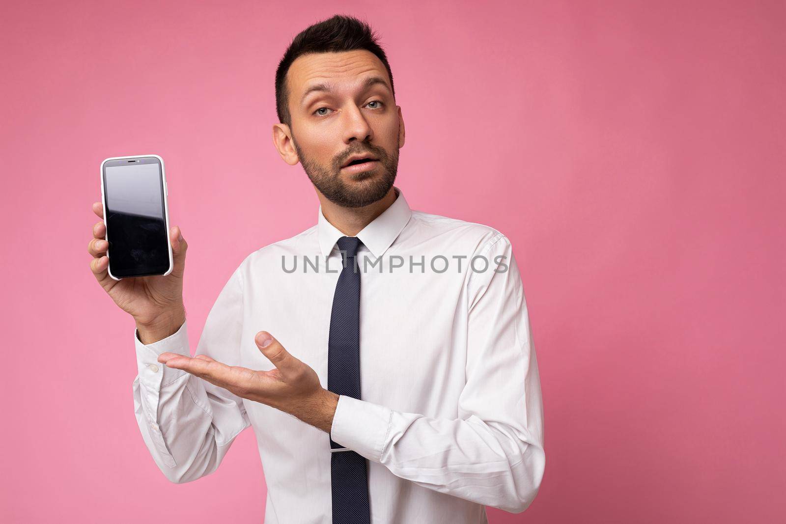 Photo of handsome smiling adult male person good looking wearing casual outfit standing isolated on background with copy space holding smartphone showing phone in hand with empty screen display for mockup pointing at gadjet looking at camera.