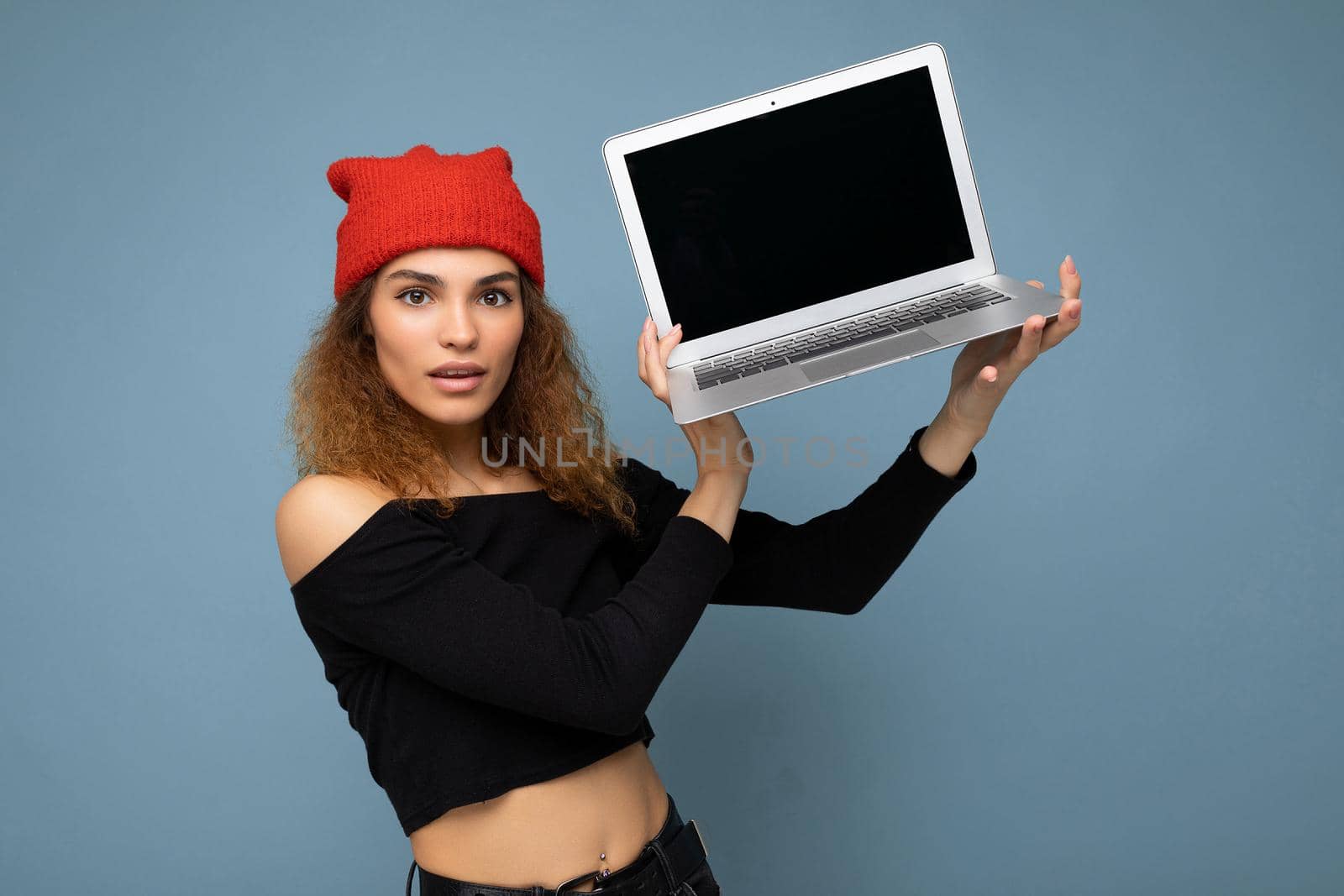 Beautiful amazed surprised shocked asking thoughtful young brunette curly woman wearing black crop top and red and orange do-rag isolated over light blue wall background holding computer laptop with empty screen monitor looking at camera by TRMK