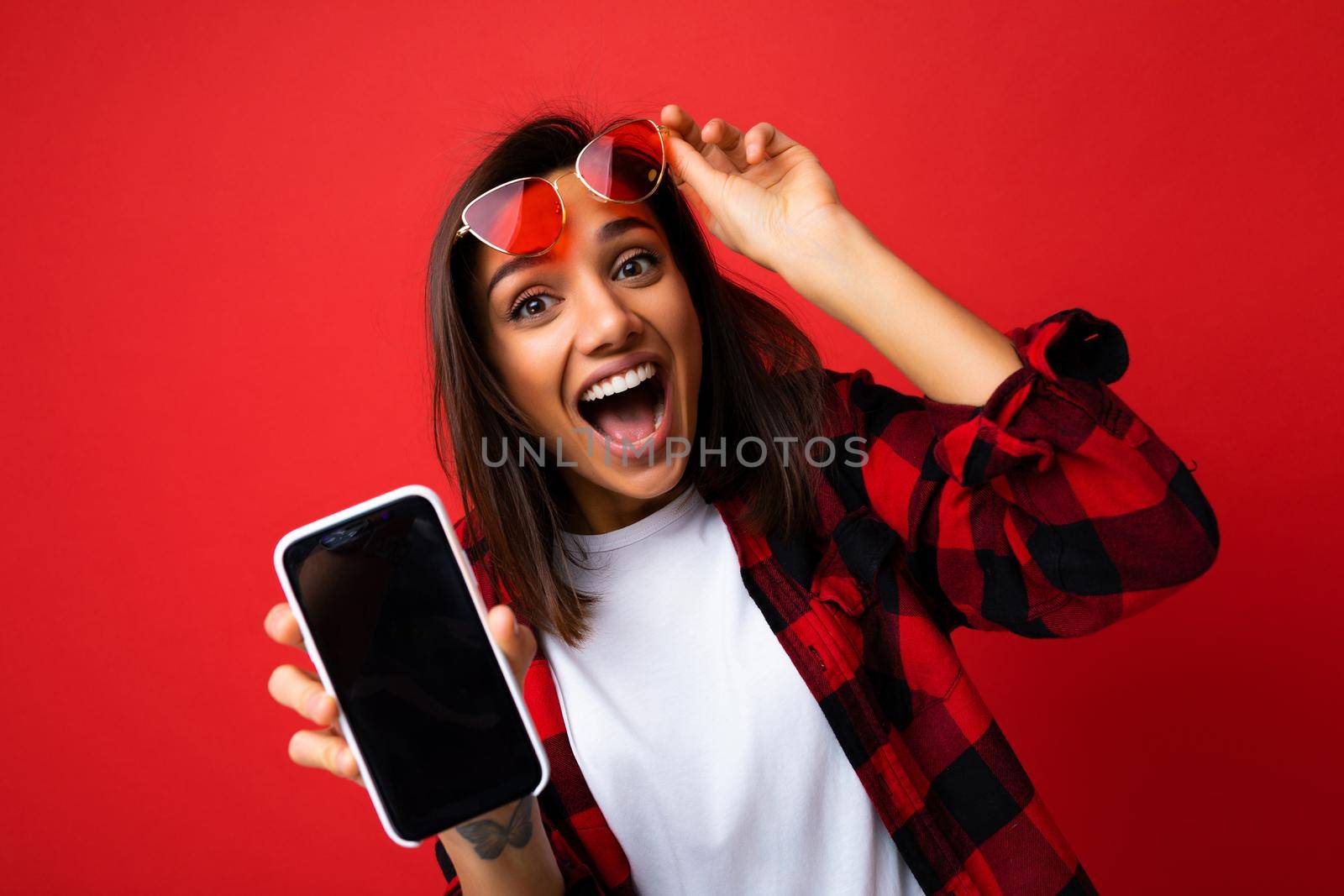 Photo of beautiful smiling surprised young woman good looking wearing casual stylish red shirt white t-shirt and red sunglasses standing isolated on red background with copy space holding smartphone showing phone in hand with empty screen display for mockup looking at camera.
