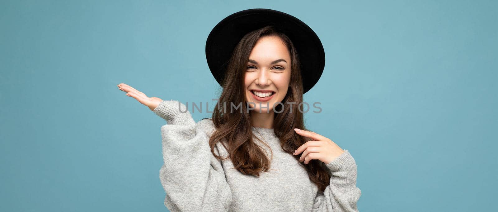 Panoramic photo of attractive brunette happy smiling young woman pointing hand at free space for text wearing grey jersey and black hat isolated on blue background.
