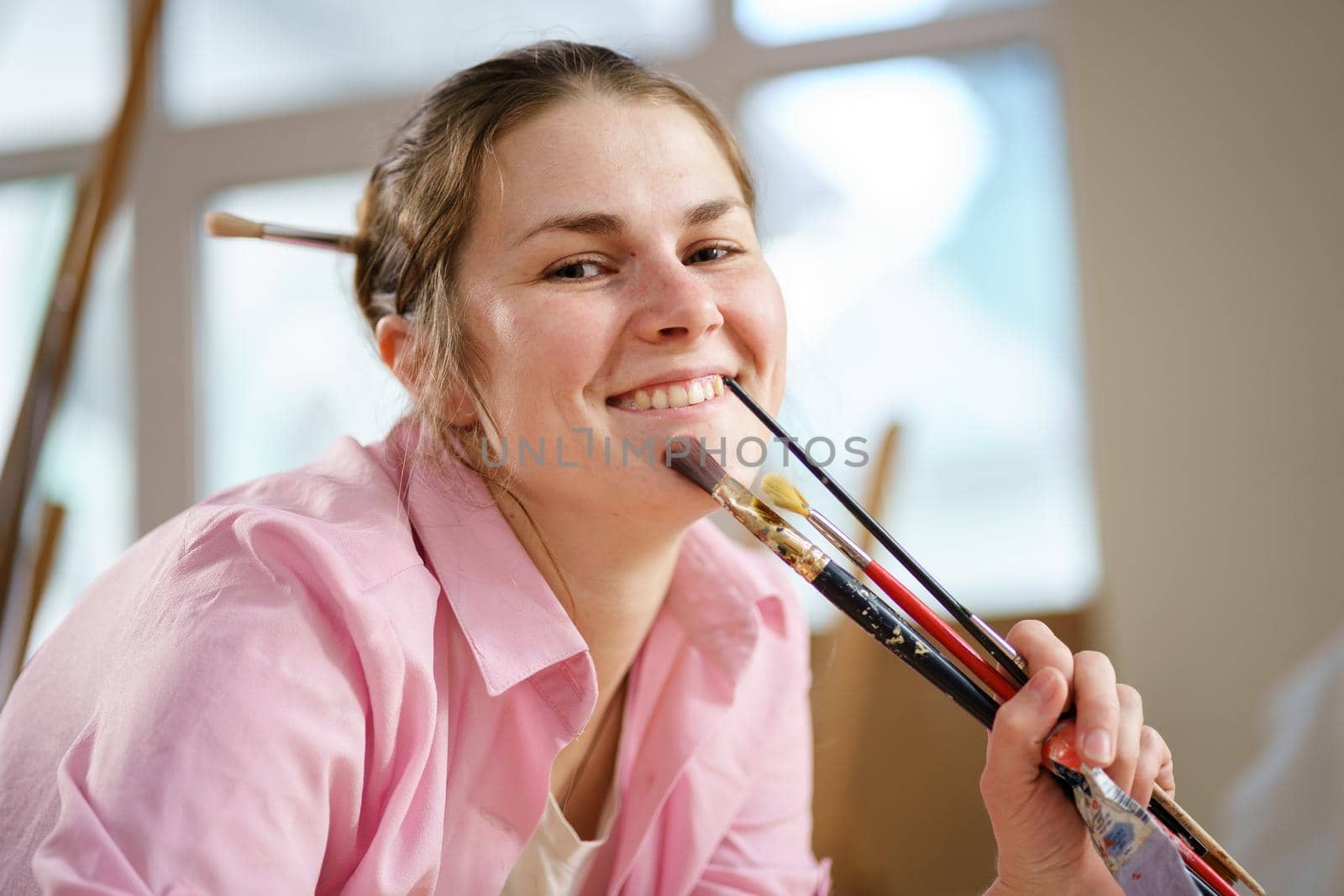 Caucasian woman artist working on a painting in bright daylight studio. Happy artist draws an art project with paints and a brush in the workshop. Hobby. Artist at work. Creative profession by Tomashevska