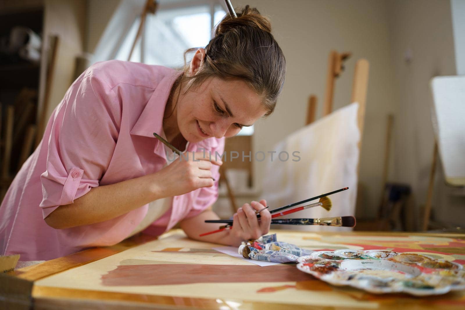 Caucasian woman artist working on a painting in bright daylight studio. Happy artist draws an art project with paints and a brush in the workshop. Hobby. Artist at work. Creative profession by Tomashevska