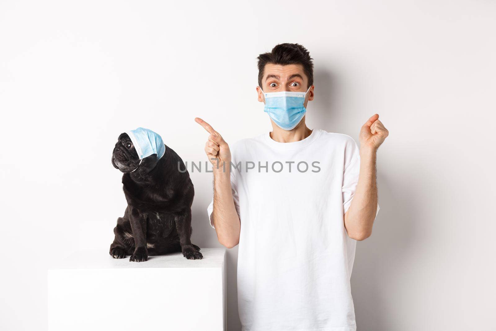 Covid-19, animals and quarantine concept. Young man and black dog wearing medical masks, pug looking at upper left corner and owner showing promo offer, rejoicing.