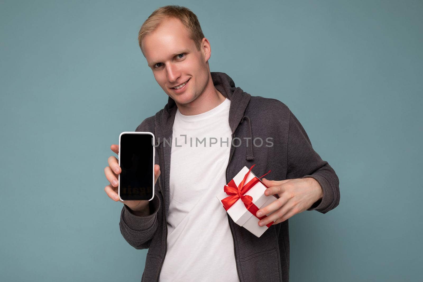 Photo of smiling cool handsome happy young man wearing grey sweater and white t-shirt standing isolated over blue background wall holding smartphone and showing phone with empty screen display and gift white box with red ribbon looking at camera.
