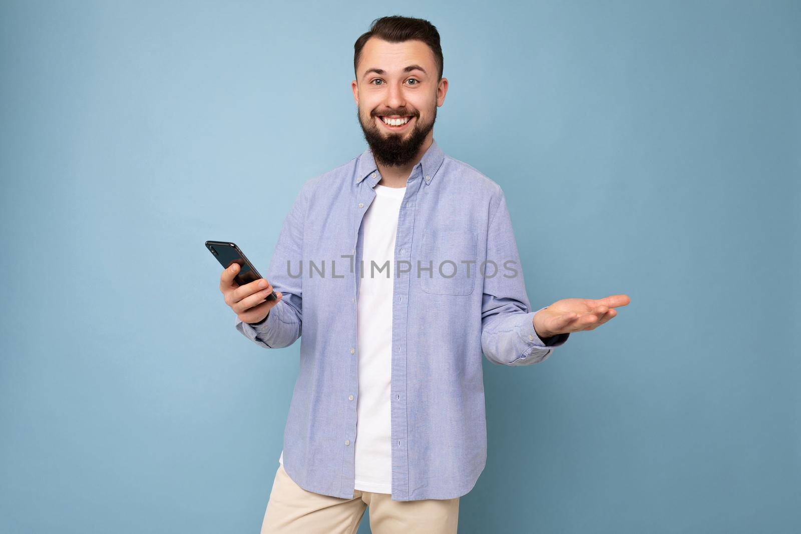 handsome young brunette unshaven man with beard wearing stylish white t-shirt and blue shirt isolated over blue background with empty space holding in hand and using phone messaging sms looking at camera and don't understanding by TRMK