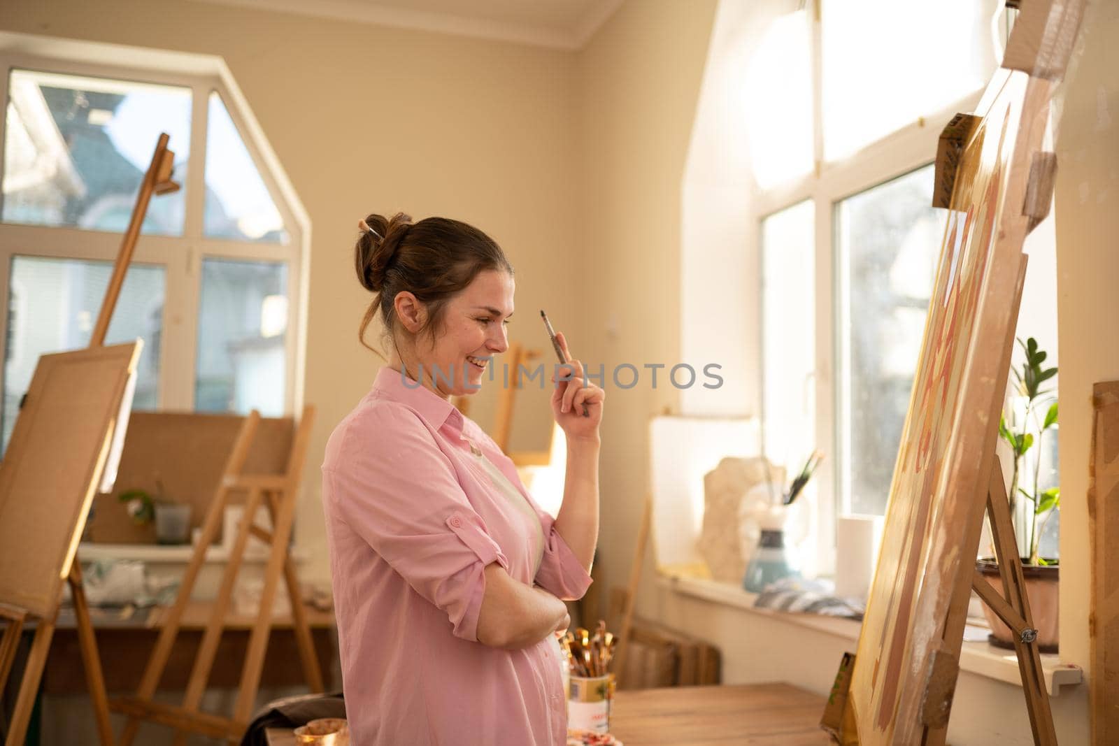 Cute woman paints on canvas in an art workshop. Artist creating picture. Art school or studio. Work with paints, brushes and easel. Hobby and leisure concept. Woman painter at workspace by Tomashevska