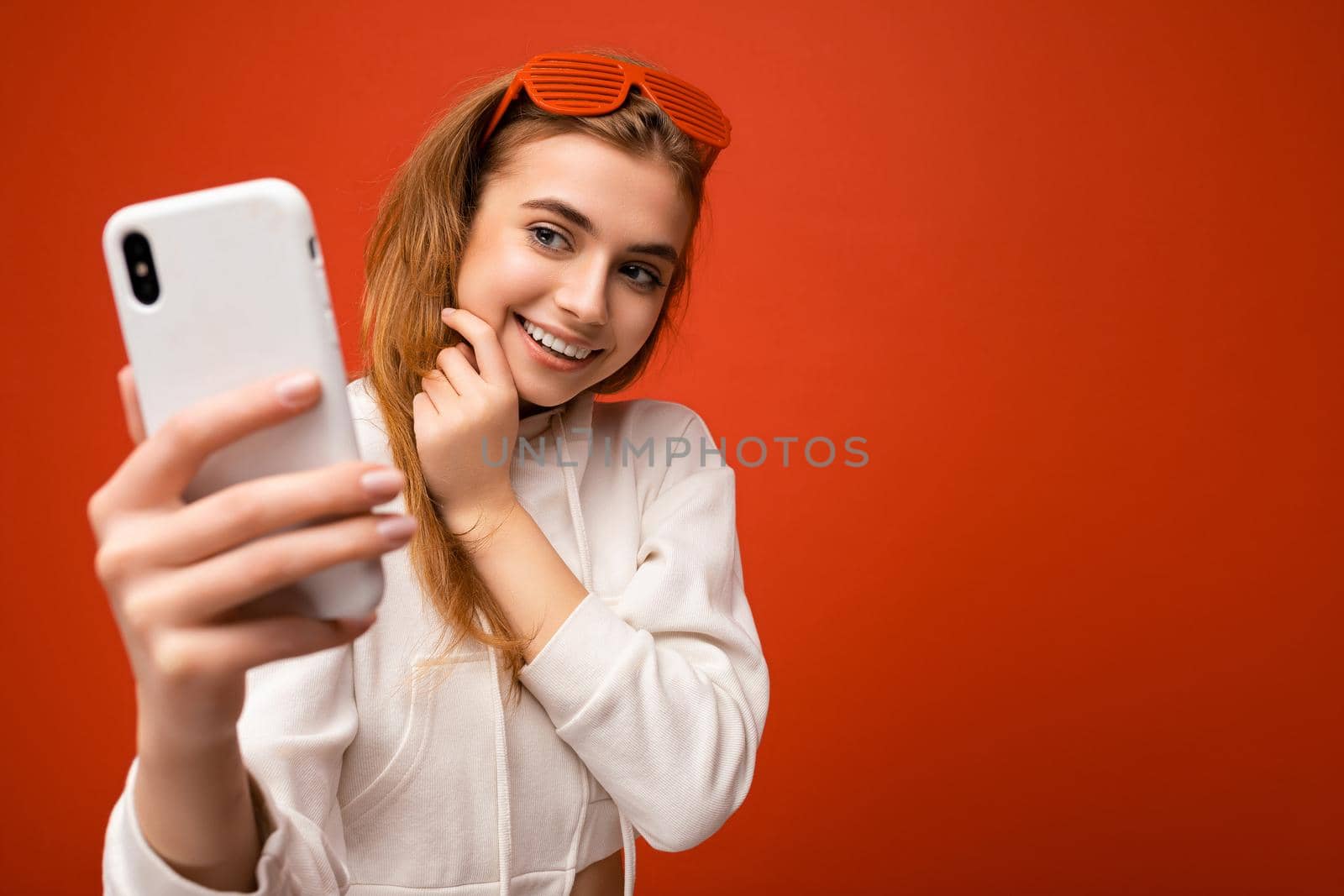 Attractive charming young smiling happy woman holding and using mobile phone taking selfie wearing stylish clothes isolated over wall background by TRMK