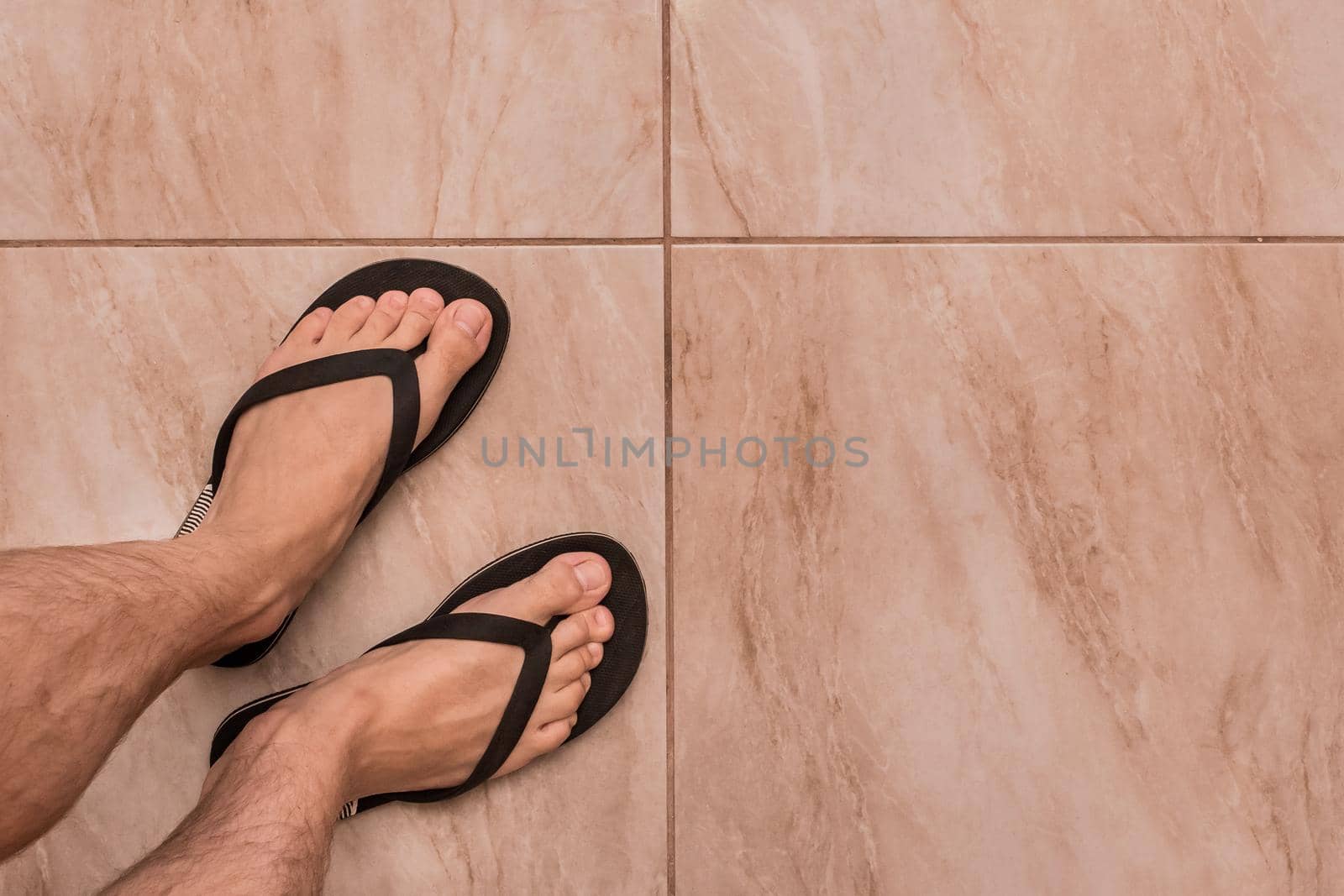 Legs of a man in black flip flops stand on a tiled floor texture background, top view by AYDO8