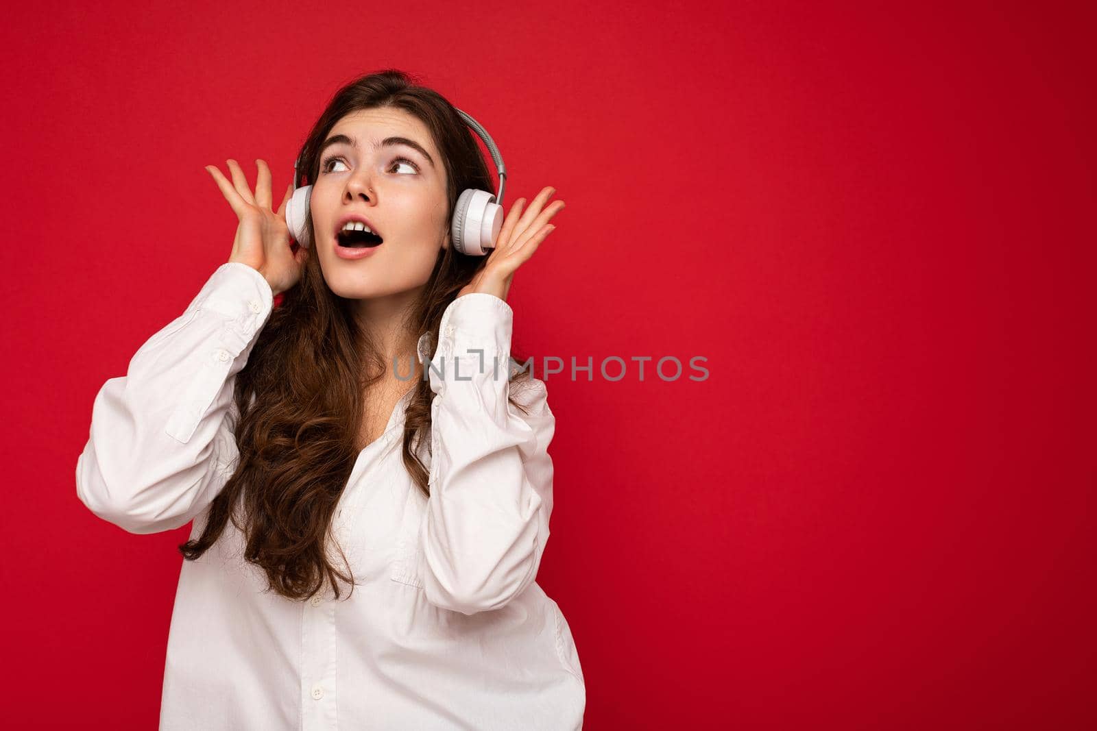 Pretty positive smiling young brunet woman wearing white shirt and optical glasses isolated over red background wearing white wireless bluetooth headphones listening to music and having fun looking to the side. empty space