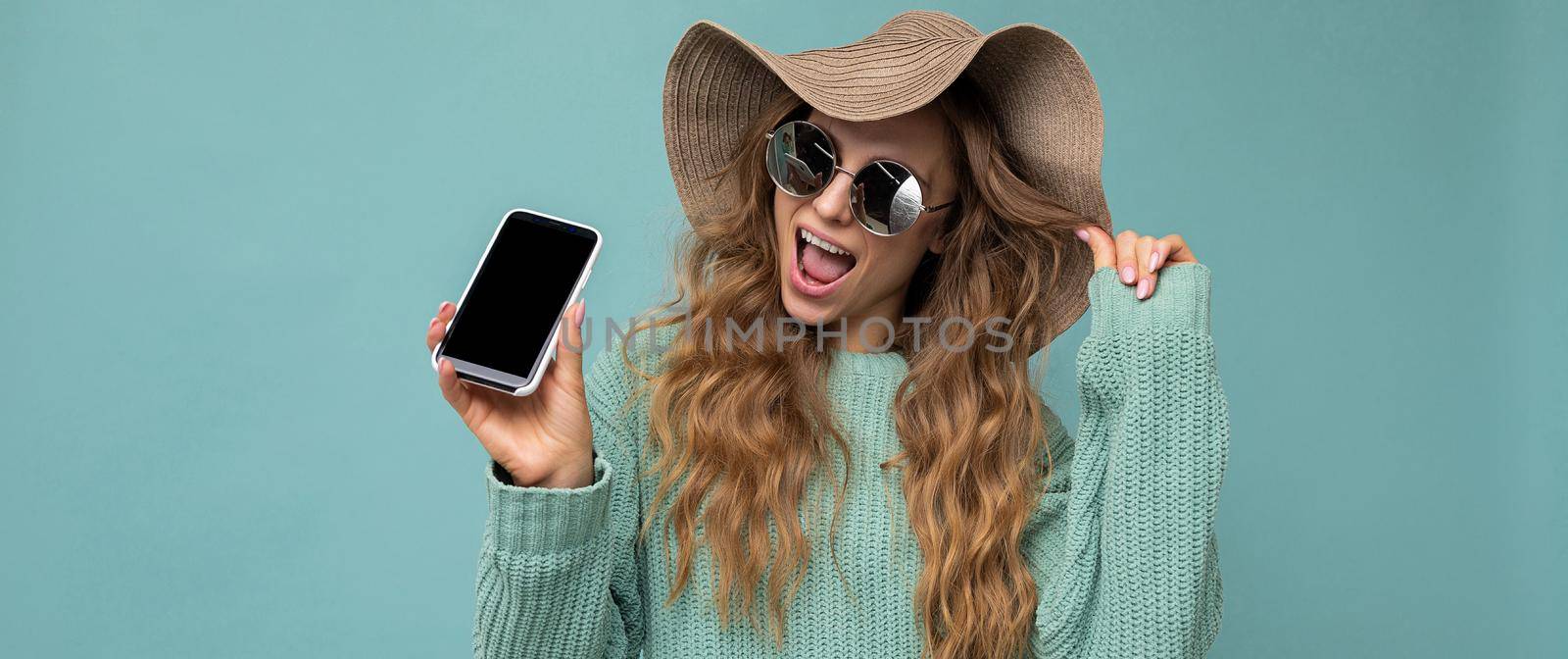 Panoramic photo of beautiful happy young blonde woman wearing sunglasses and summer hat isolated on blue background with copy space holding smartphone showing phone in hand with empty screen display looking at camera with open mouth. Mockup
