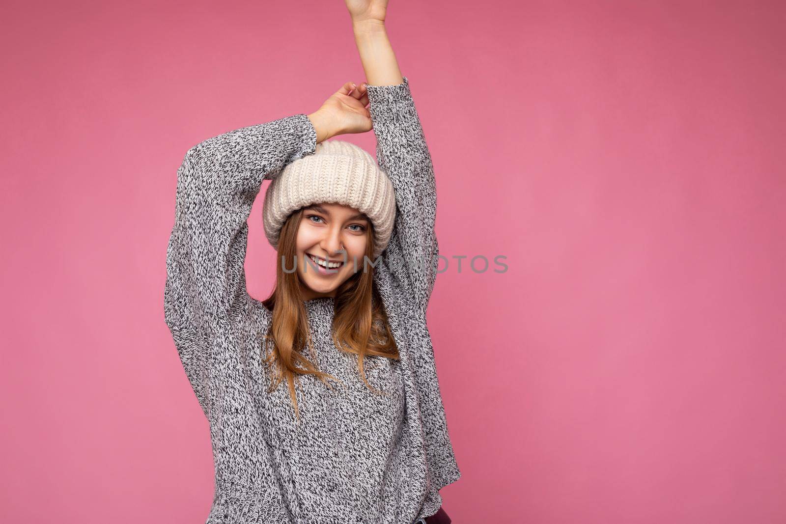 Shot of attractive happy funny amazing young dark blond woman standing isolated over pink background wall wearing grey sweater and beige hat looking at camera. Empty space