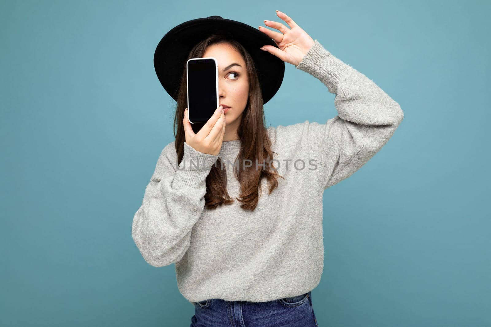 woman wearing black hat and grey sweater holding mobilephone showing smartphone isolated on background looking to the side. mock up