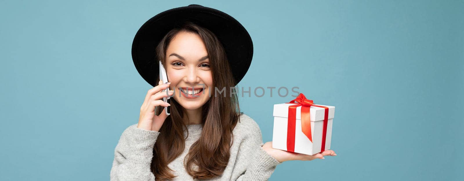 Shot of pretty smiling positive young brunette woman isolated over blue background wall wearing stylish black hat and grey sweater holding gift box talking on smartphone and looking at camera.