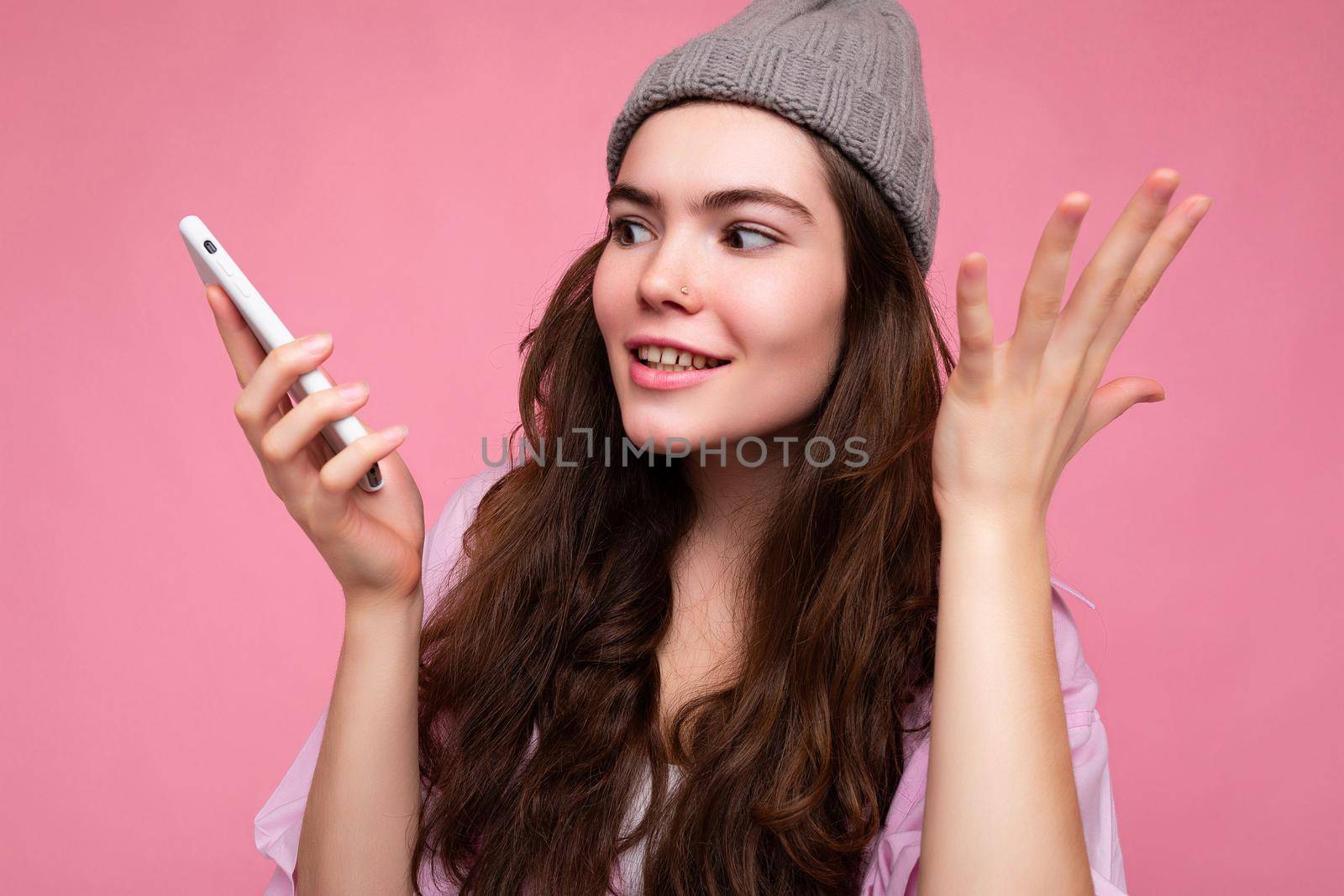 Closeup photo of Emotional attractive young brunette woman wearing stylish pink shirt and grey hat isolated over pink background holding in hand and using mobile phone communicating and recording voice message.