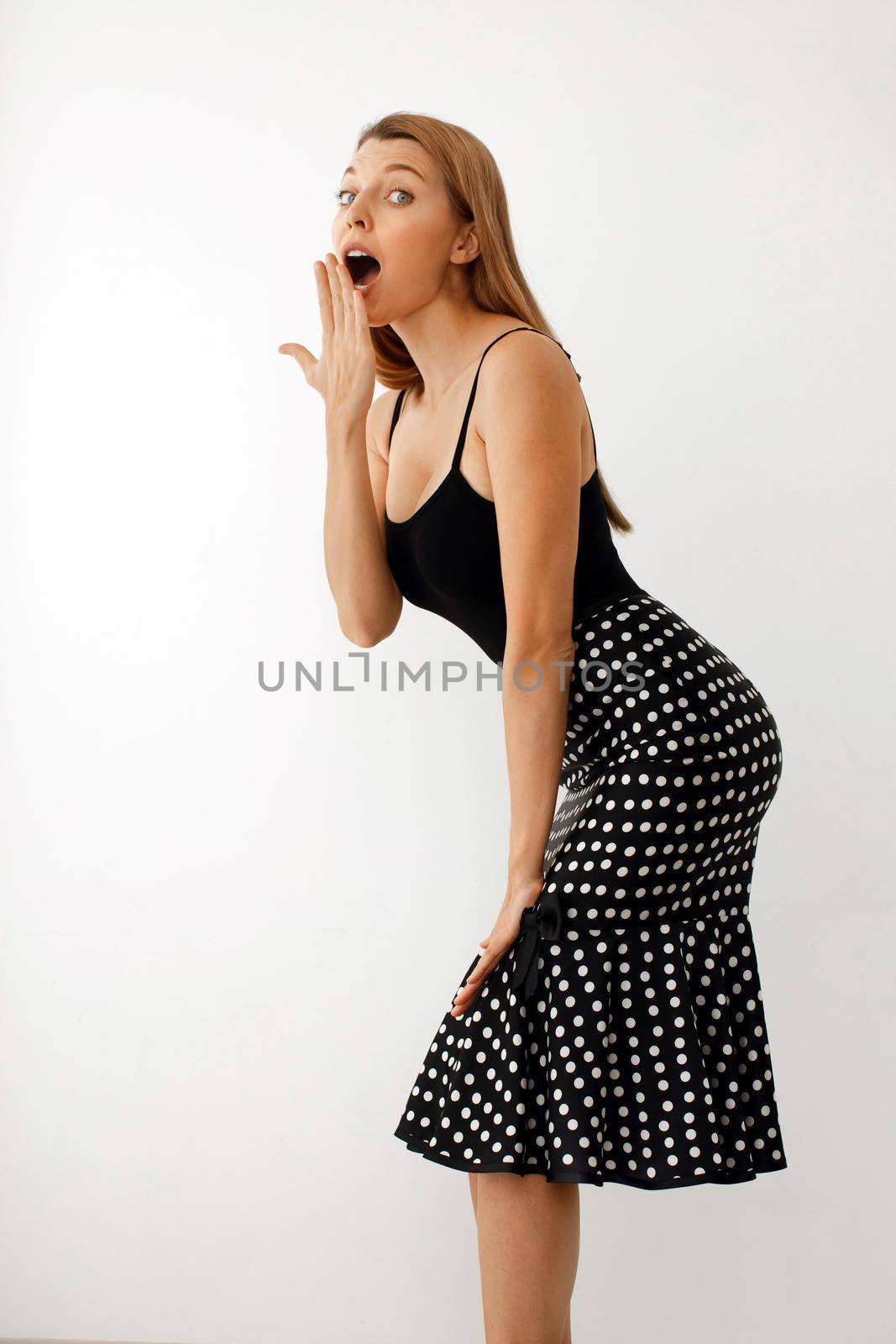 Side view of surprised young long haired female in black top and polka dot skirt covering open mouth with hand while gossiping