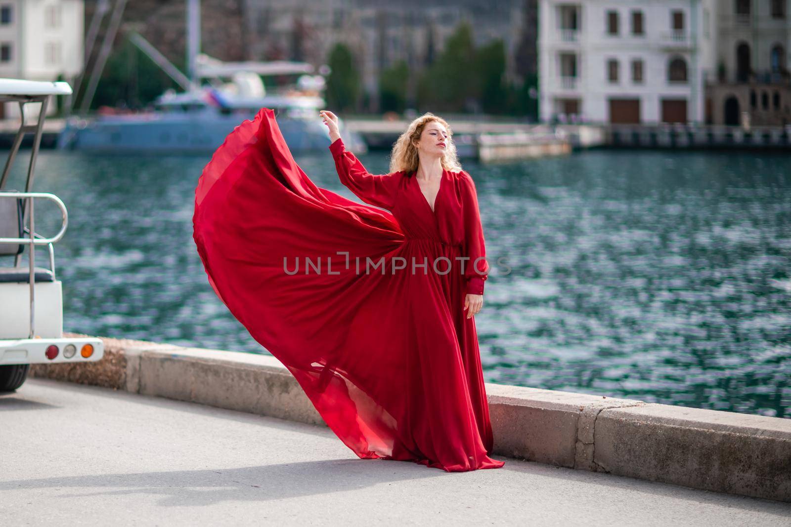 A woman in a red dress, a fashion model with long silk wings in a flowing dress, flying fabric on the embankment