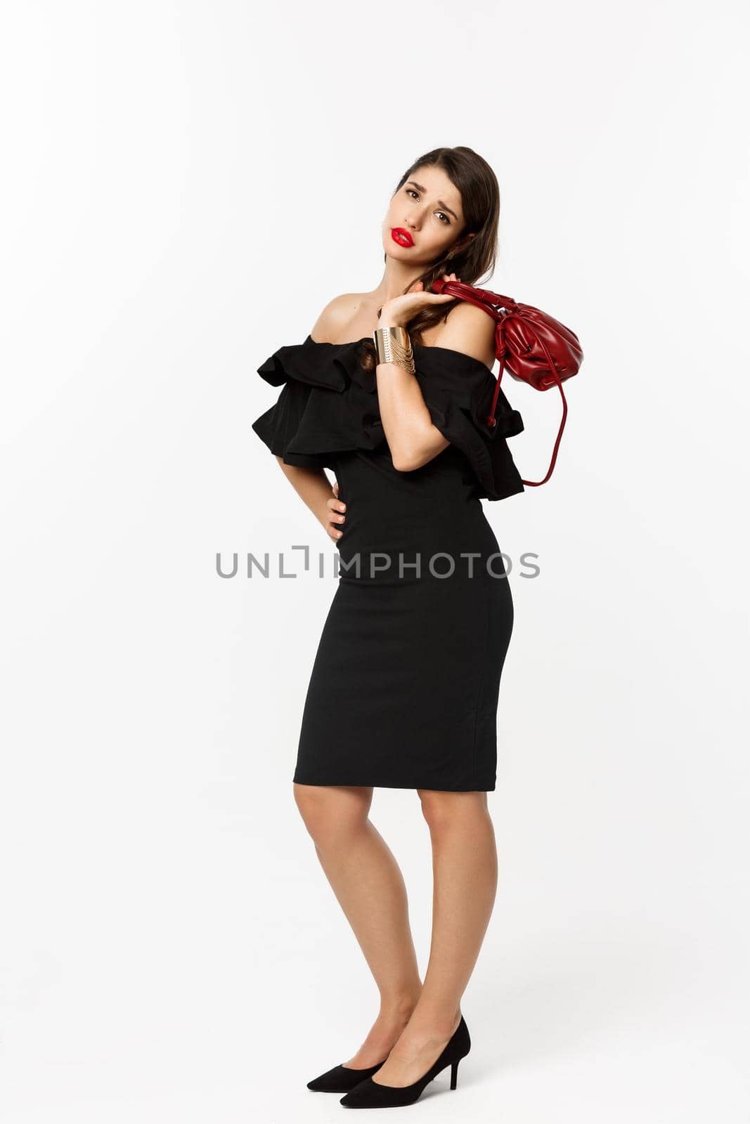 Beauty and fashion concept. Full length of tired young woman in high heels and elegant dress, holding purse on shoulder and looking with fatigue at camera, white background.