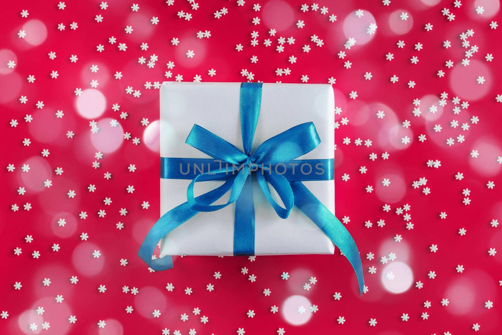 Gift box wrapped in white paper with a blue bow on festive crimson background with many snowflakes. Copyspace for your text. Flat lay style. Christmas, New Year or birthday celebration concept