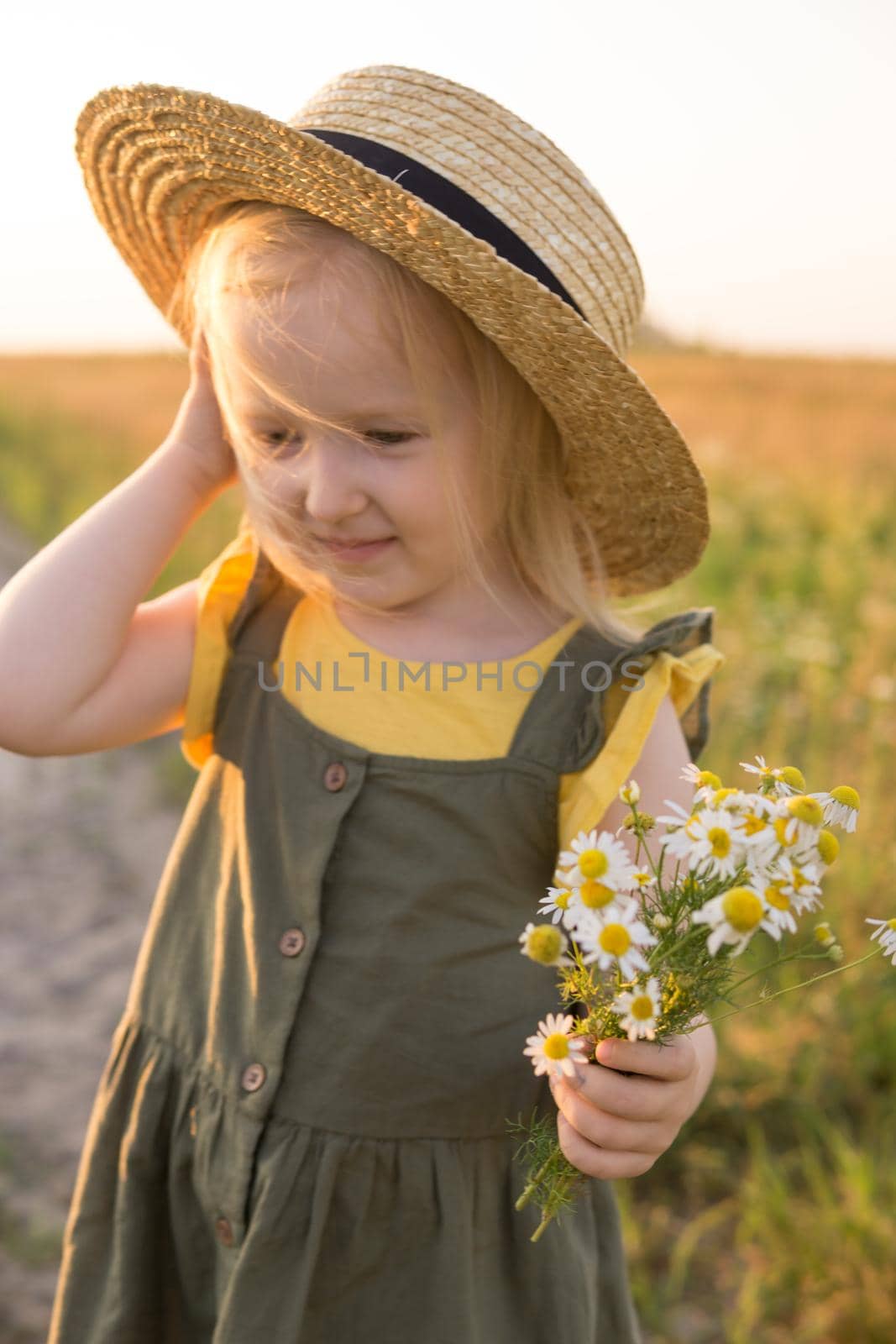 A little blonde girl in a straw hat walks in a field with a bouquet of daisies. The concept of walking in nature, freedom and an eco-friendly lifestyle.