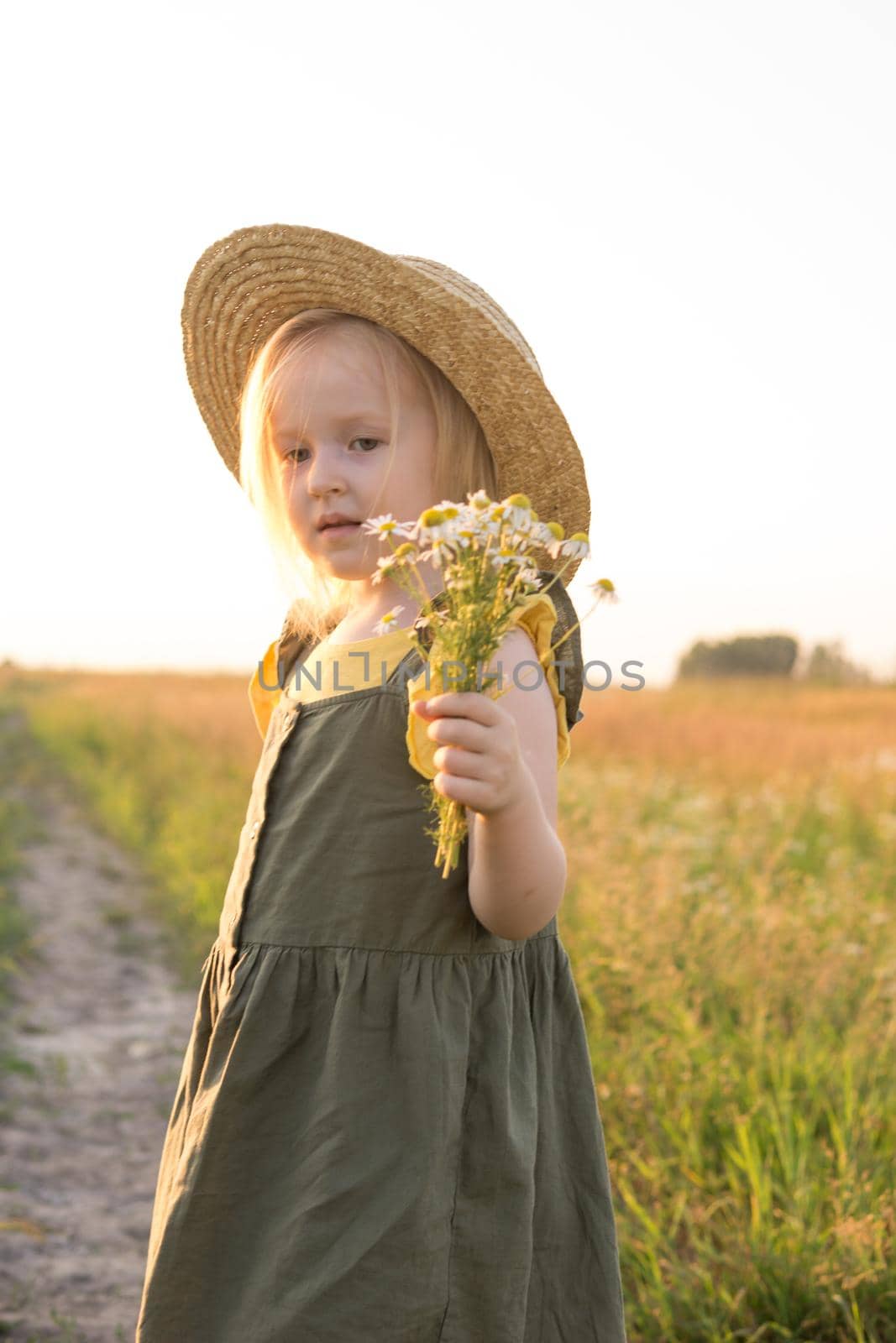 A little blonde girl in a straw hat walks in a field with a bouquet of daisies. The concept of walking in nature, freedom and an eco-friendly lifestyle.