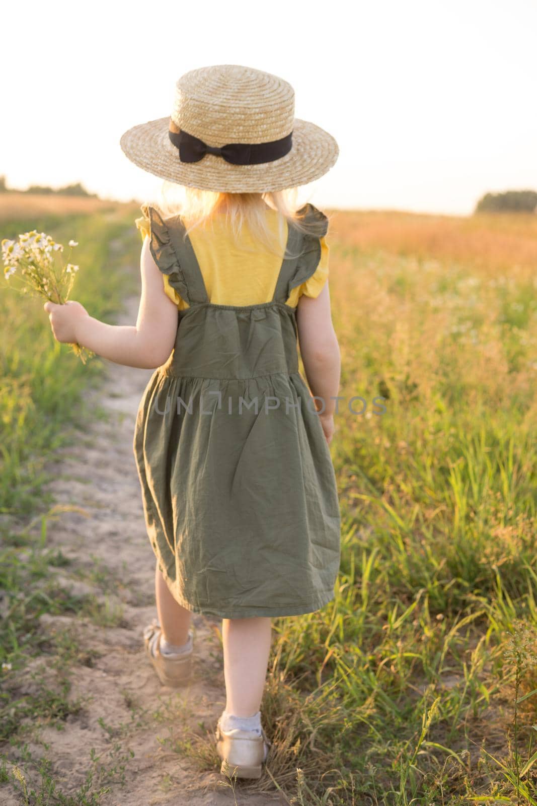 A little blonde girl in a straw hat walks in a field with a bouquet of daisies. The concept of walking in nature, freedom and an eco-friendly lifestyle.