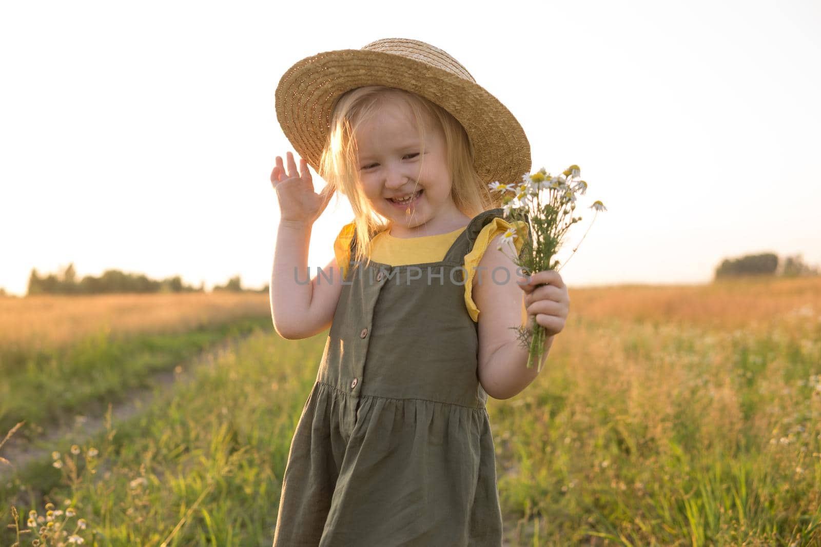 A little blonde girl in a straw hat walks in a field with a bouquet of daisies. The concept of walking in nature, freedom and an eco-friendly lifestyle.