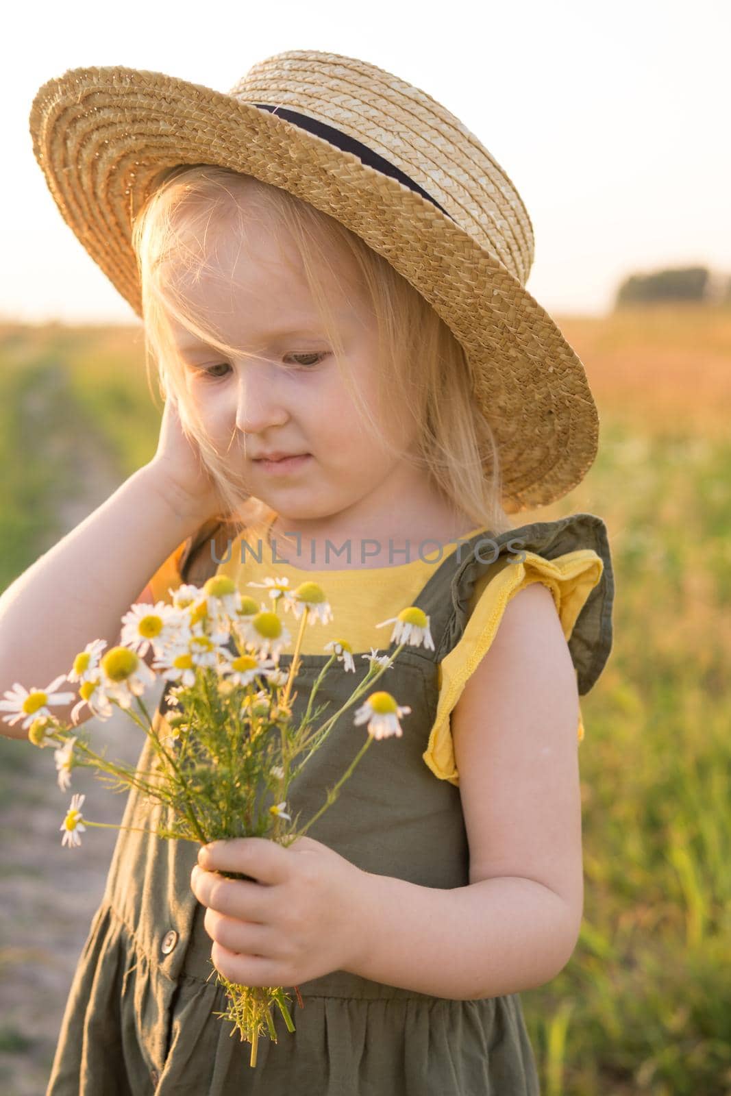 A little blonde girl in a straw hat walks in a field with a bouquet of daisies. The concept of walking in nature, freedom and an eco-friendly lifestyle.