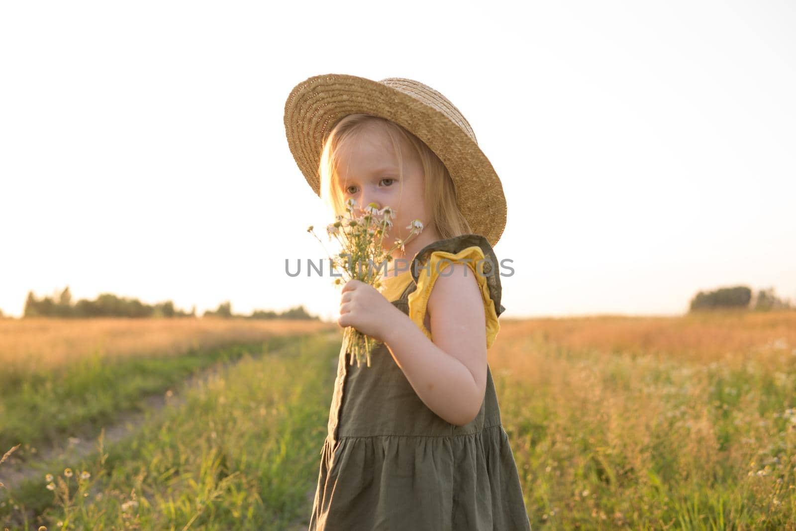 A little blonde girl in a straw hat walks in a field with a bouquet of daisies. The concept of walking in nature, freedom and an eco-friendly lifestyle.