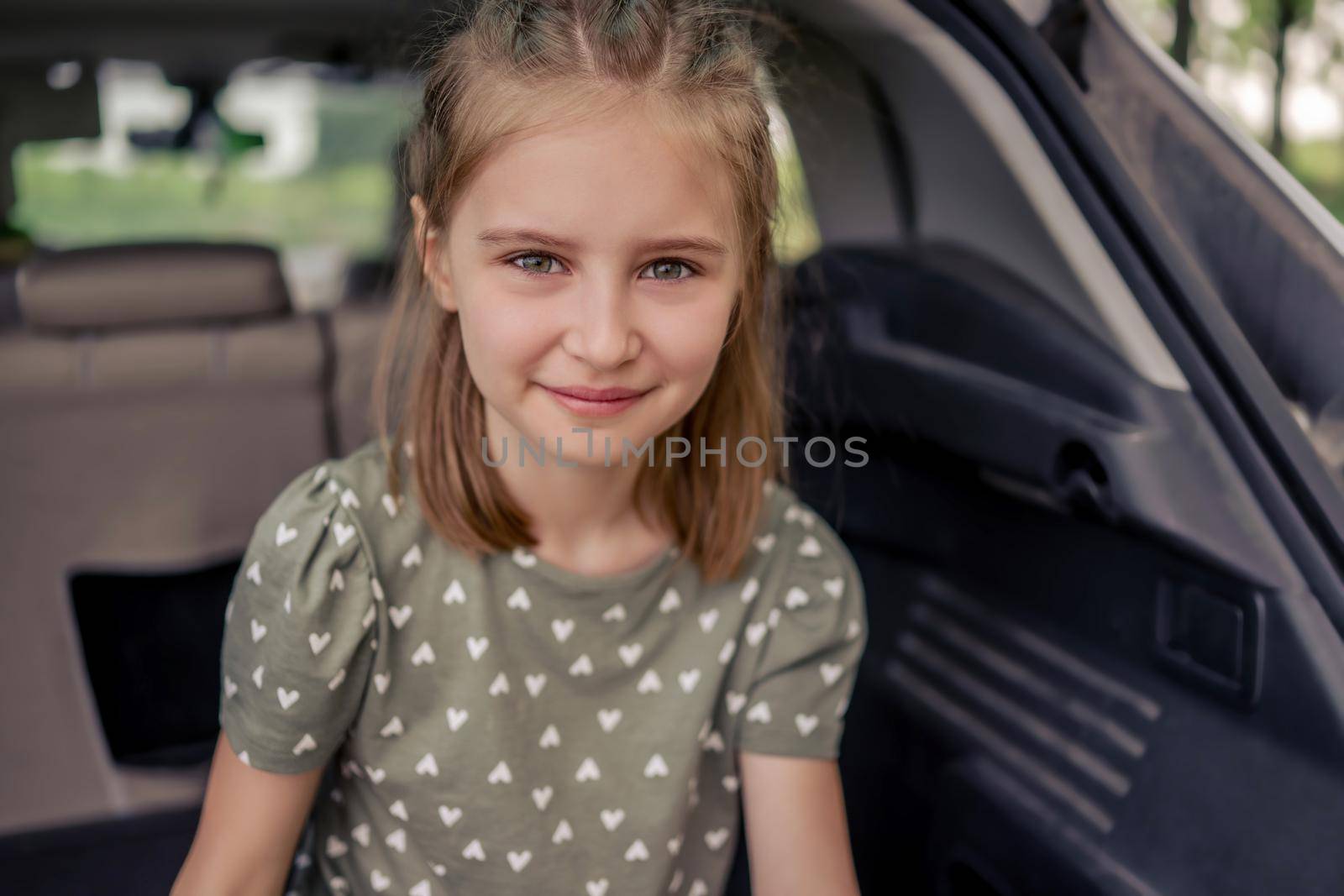 Closeup portrait of cute preteen girl with beautiful hairstyle sitting in the car trunk and smiling looking at the camera. Happy child kid in the vehicle at the nature during summer trip