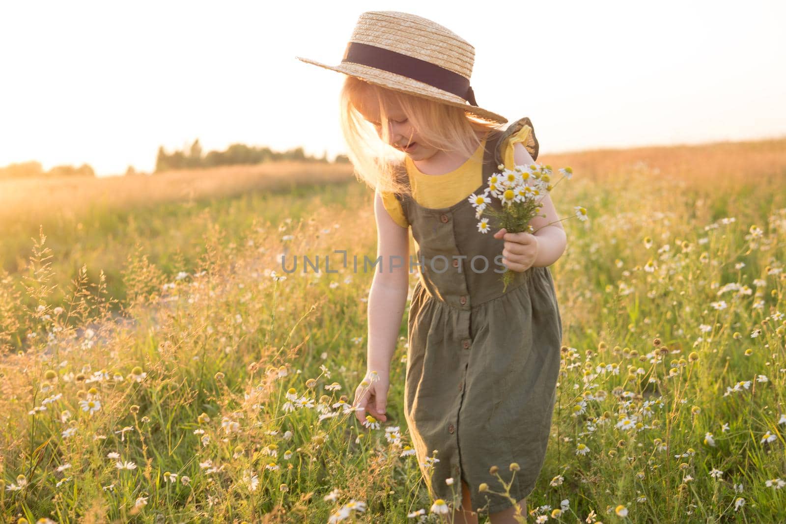 A little blonde girl in a straw hat walks in a field with a bouquet of daisies. The concept of walking in nature, freedom and an eco-friendly lifestyle.