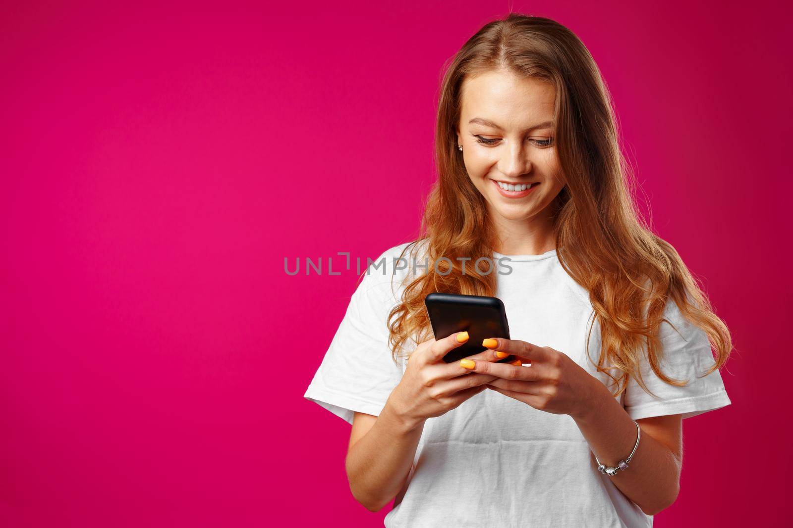 Young smiling woman standing and using her smartphone against pink background