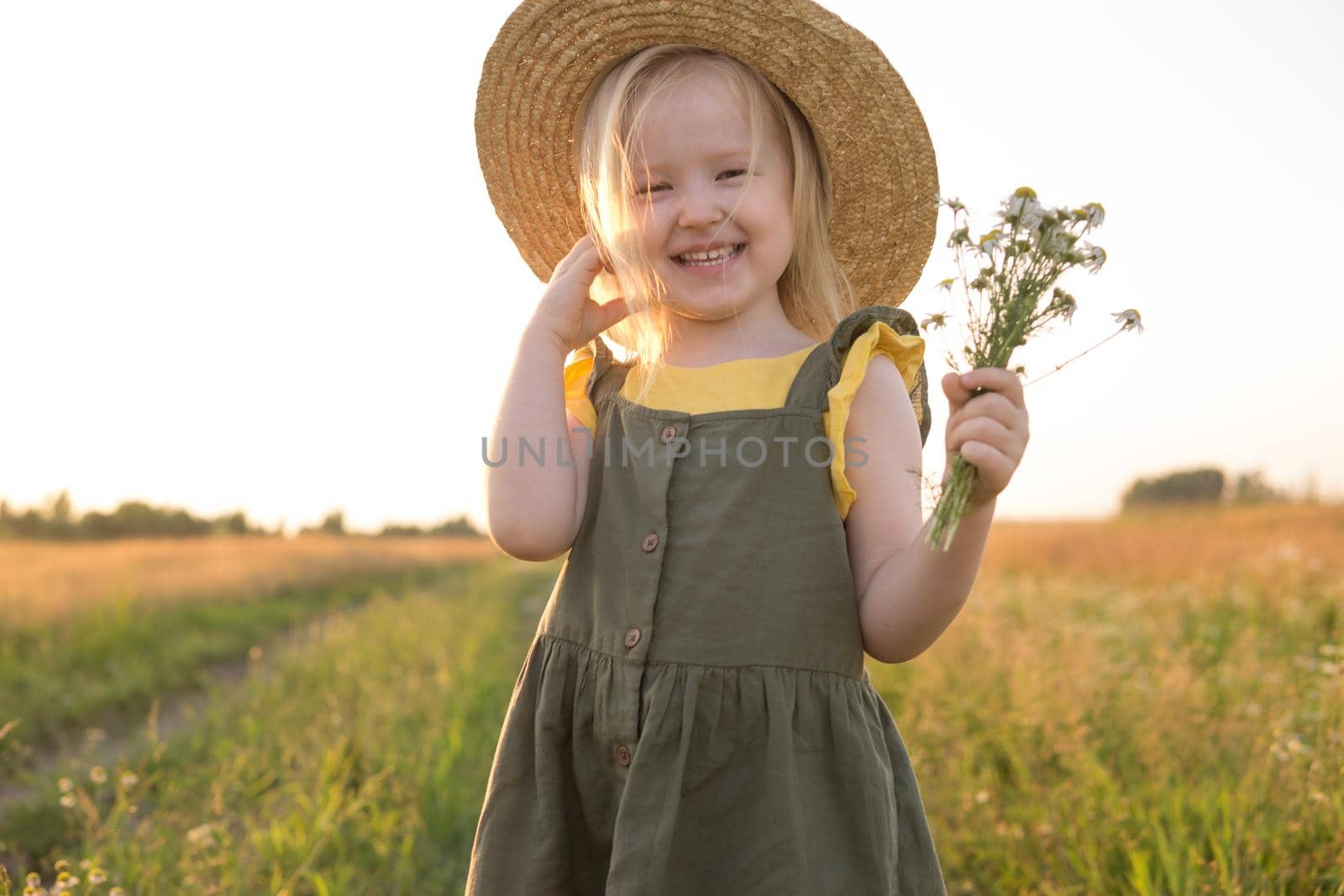 A little blonde girl in a straw hat walks in a field with a bouquet of daisies. The concept of walking in nature, freedom and an eco-friendly lifestyle.