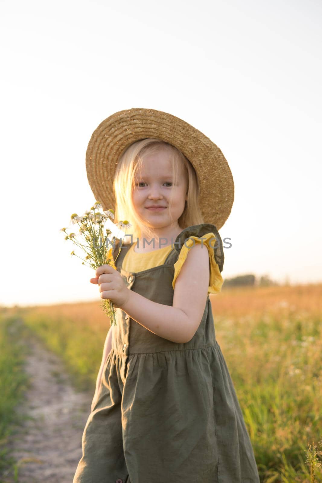 A little blonde girl in a straw hat walks in a field with a bouquet of daisies. The concept of walking in nature, freedom and an eco-friendly lifestyle.