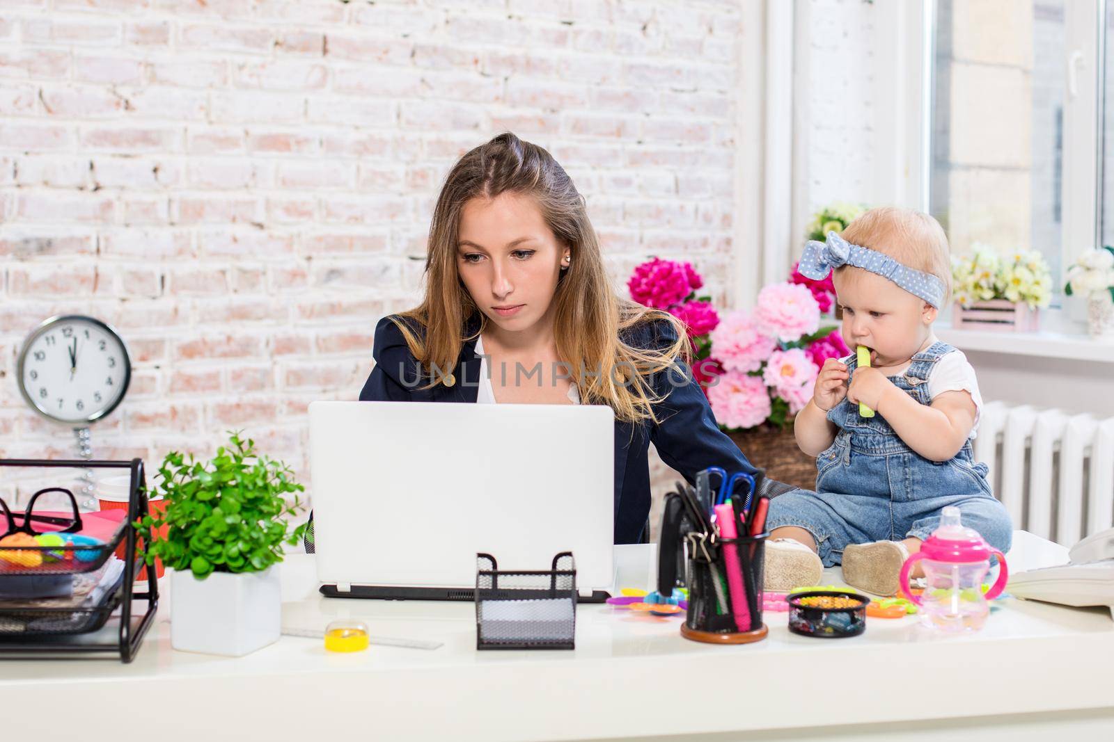 Cheerful young beautiful businesswoman looking at laptop while sitting at her working place with her little daughter by nazarovsergey