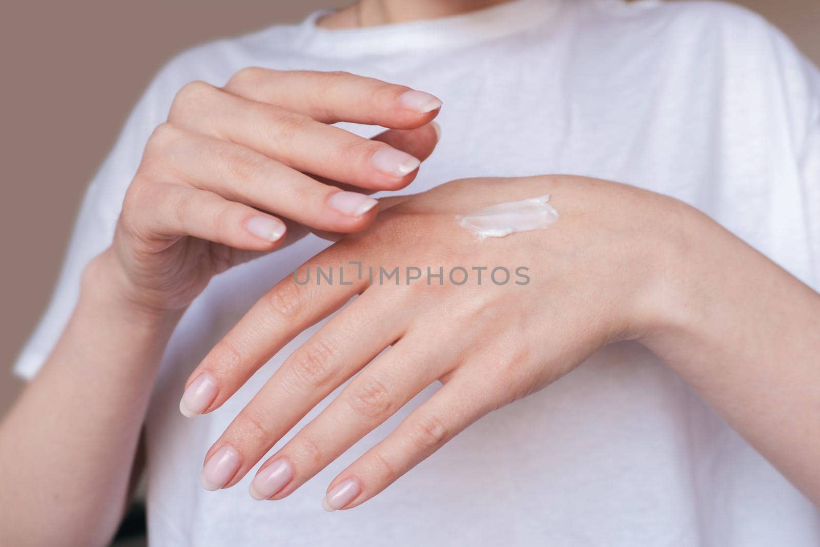 Female hands with natural pink manicure applying moisturizing cream or lotion on beige background. Bodycare, skin protection concept. Woman in white uses beauty product. Spa procedure closeup.