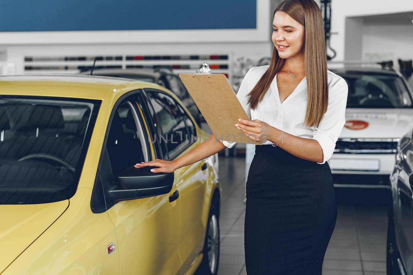 Attractive young female car dealer standing in showroom near a new car