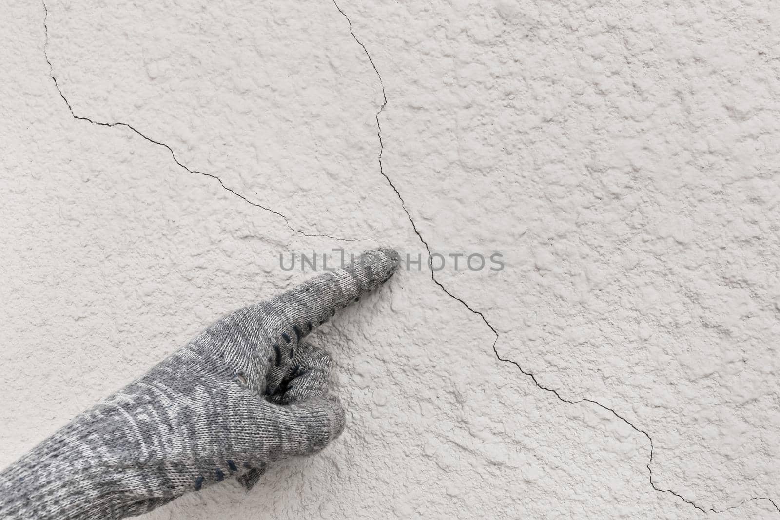 Hand of industrial worker in construction glove points to cracks on white plaster wall texture background.