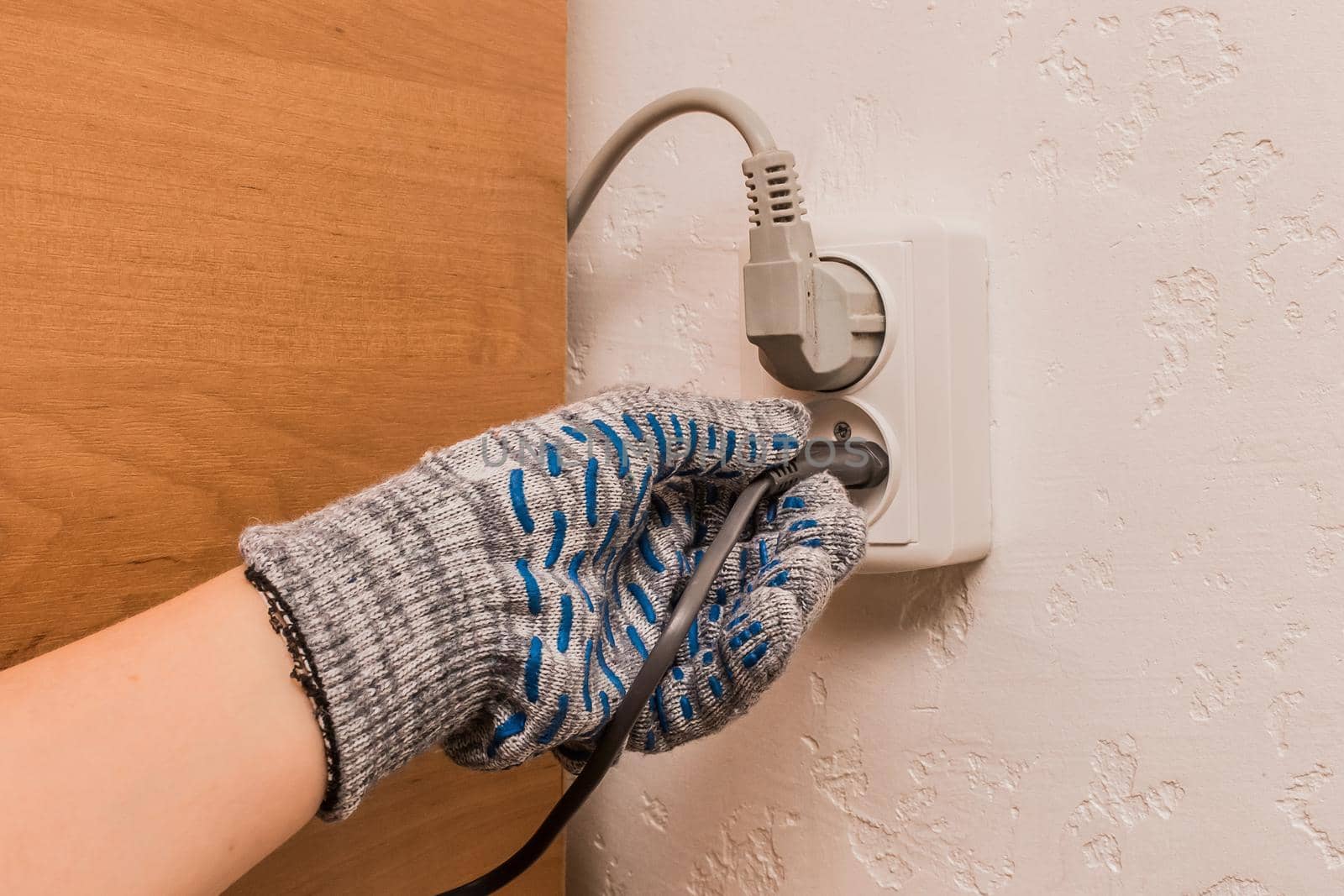 The hand of a male construction worker in protective gloves connects a plug to a double socket against the background of a wall.