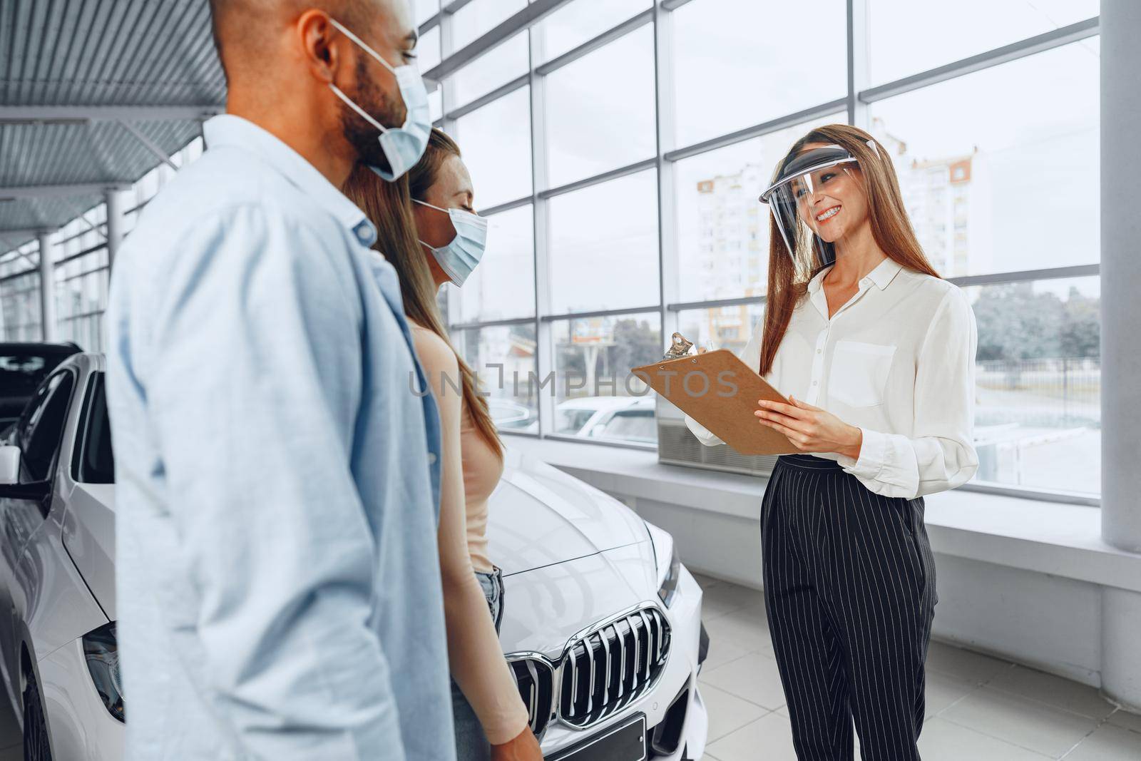 Woman car dealer consulting buyers wearing medical face shield. Coronavirus job requirements concept