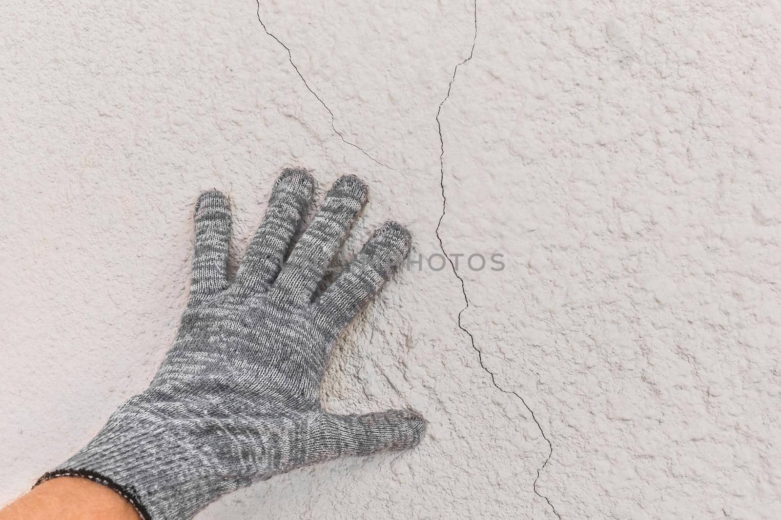 Hand of a working man in a construction glove examines cracks on a white wall background. Repair concept by AYDO8