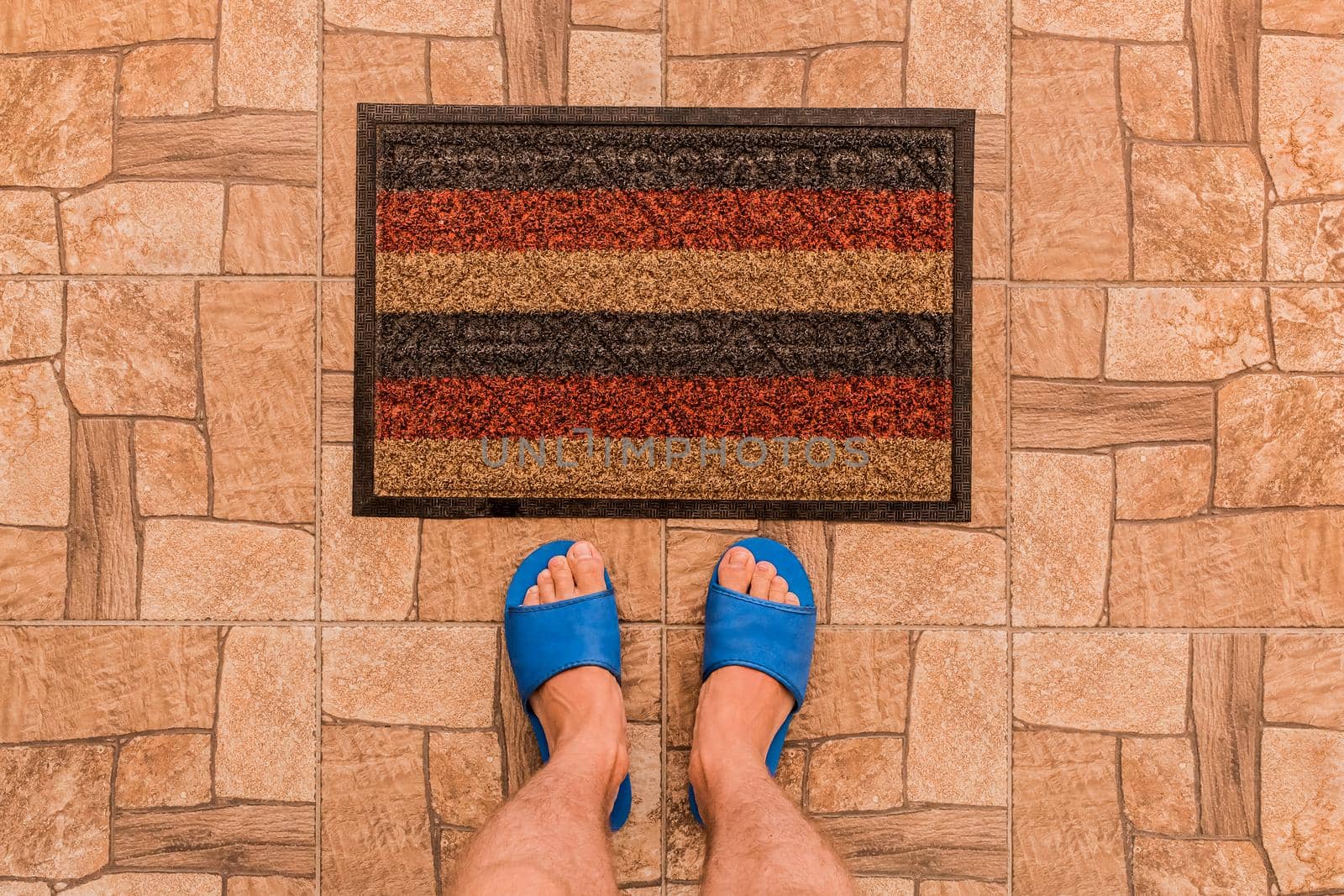 Male feet in blue house slippers stand in front of a foot mat on a brown tiled floor texture background, top view by AYDO8