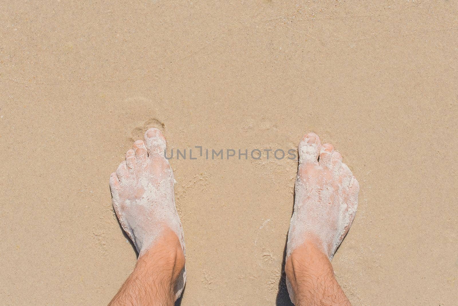 The young man's legs stand on the beach sand and are washed by blue sea water, top view.